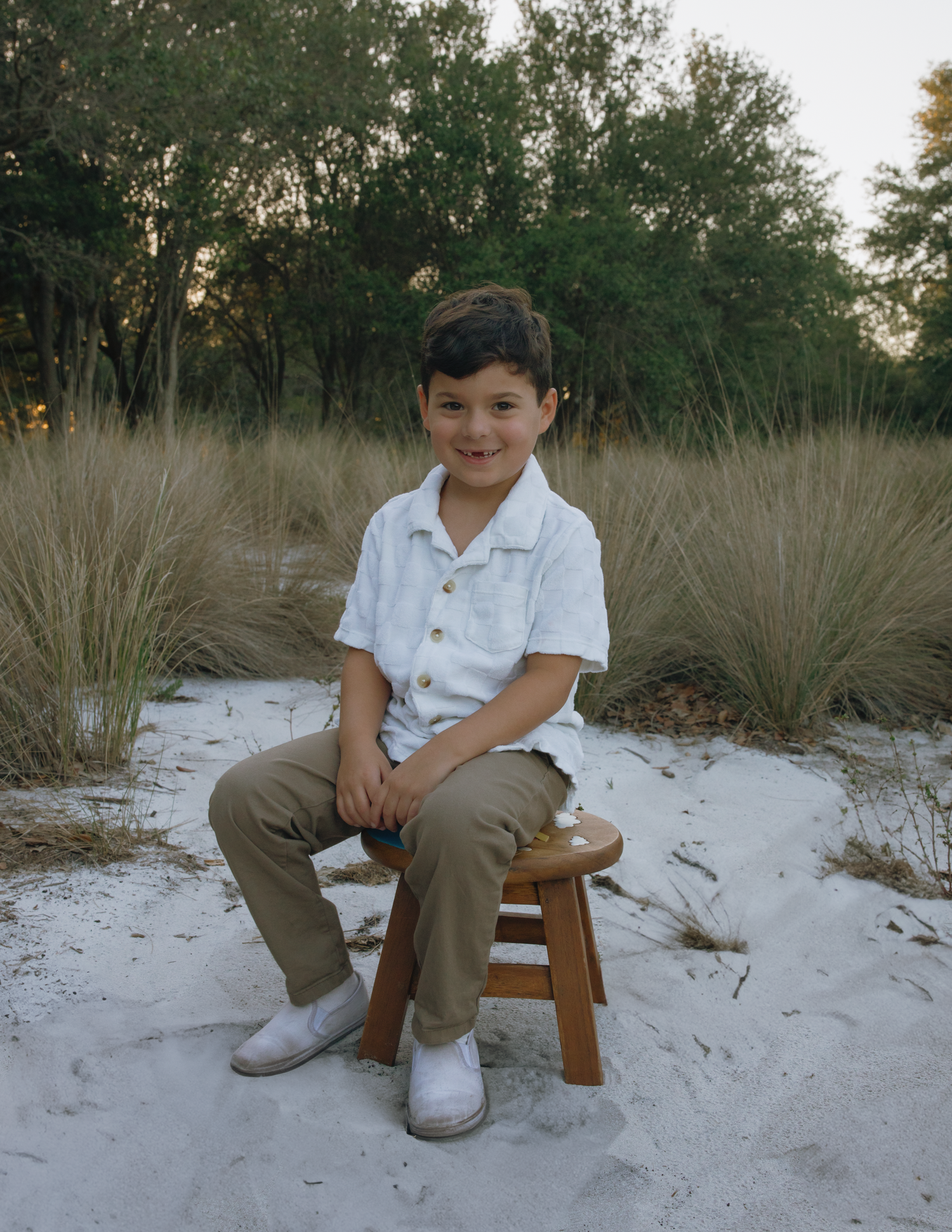 A young person sits on a wooden stool against a backdrop of tall grasses and trees.