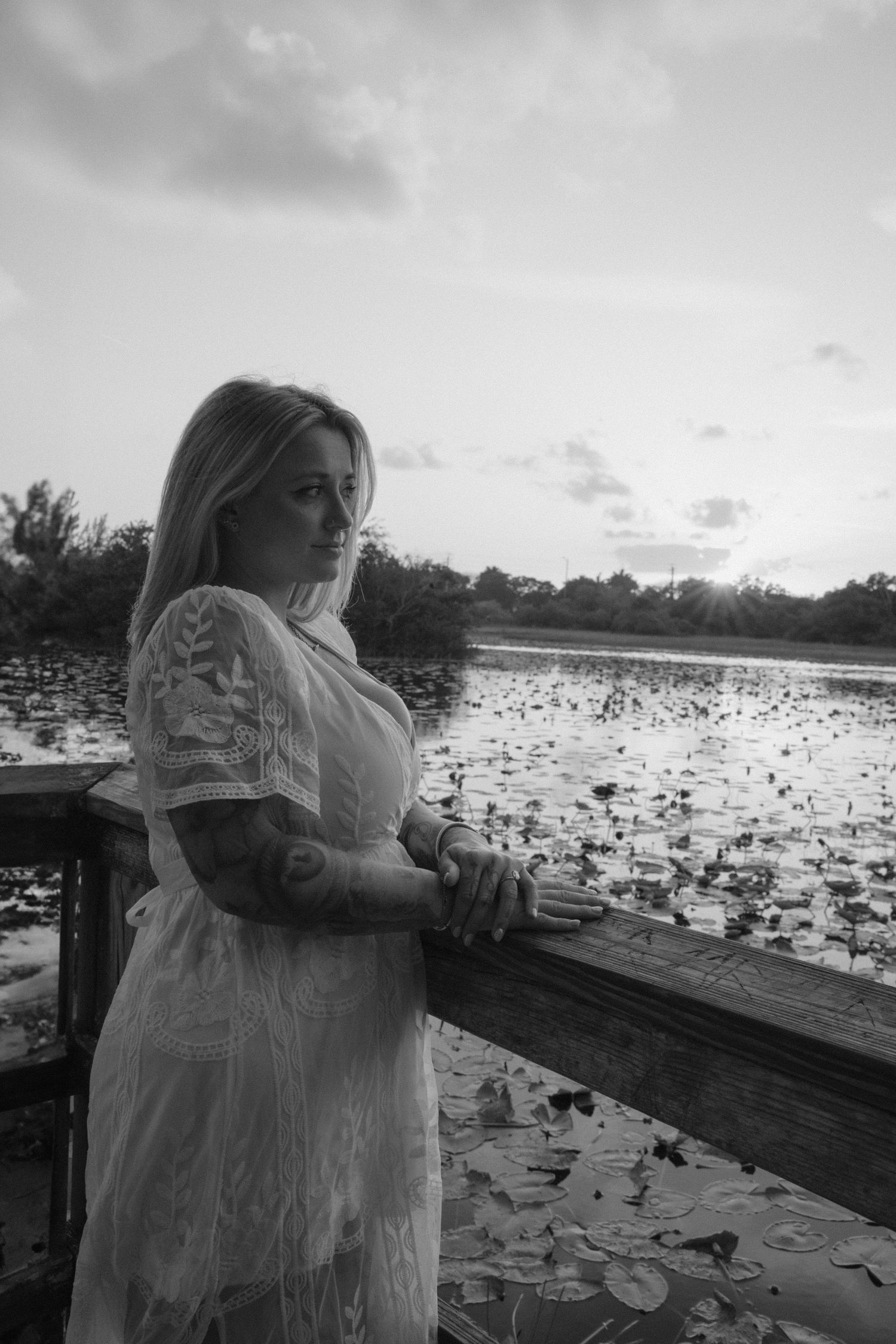 Person in white lace dress standing at a lakeside boardwalk at sunset.