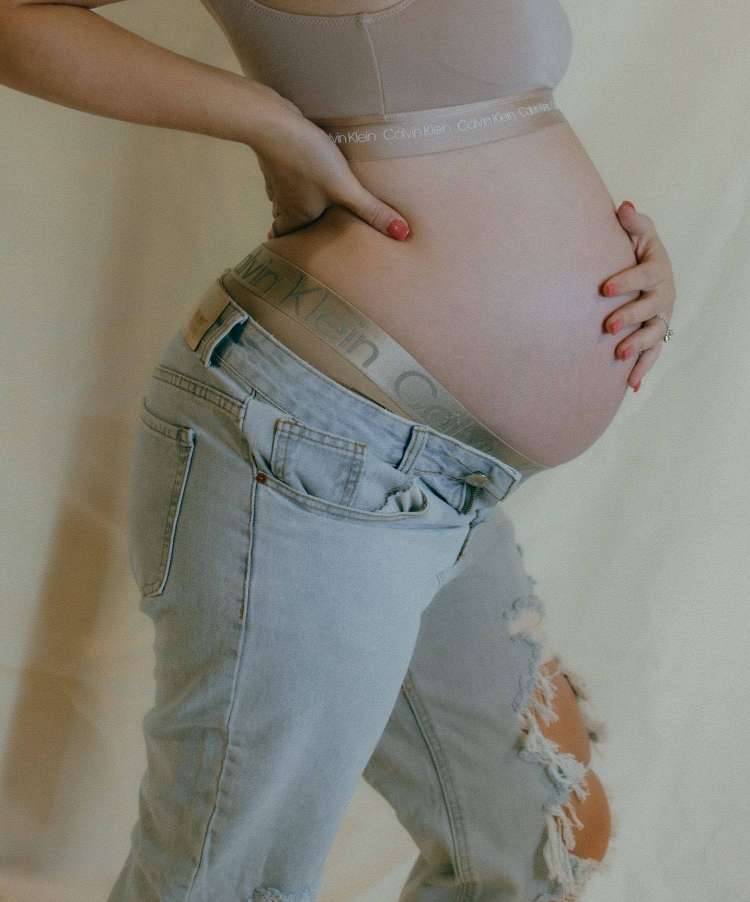 Pregnant woman in distressed jeans showing baby bump against neutral background.