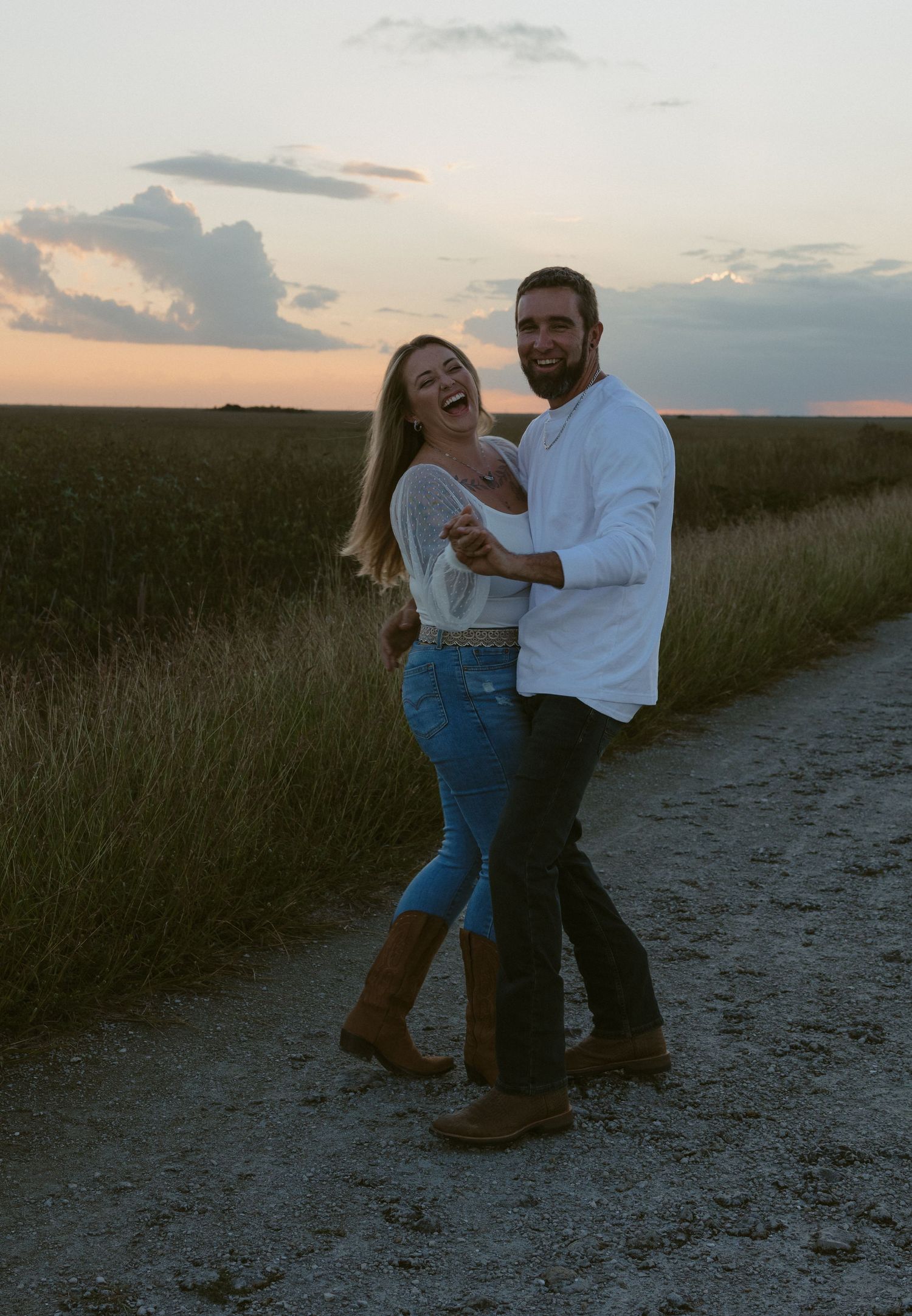 A laughing couple embraces on a rural dirt path at sunset, surrounded by green fields under a dramatic cloudy sky.
