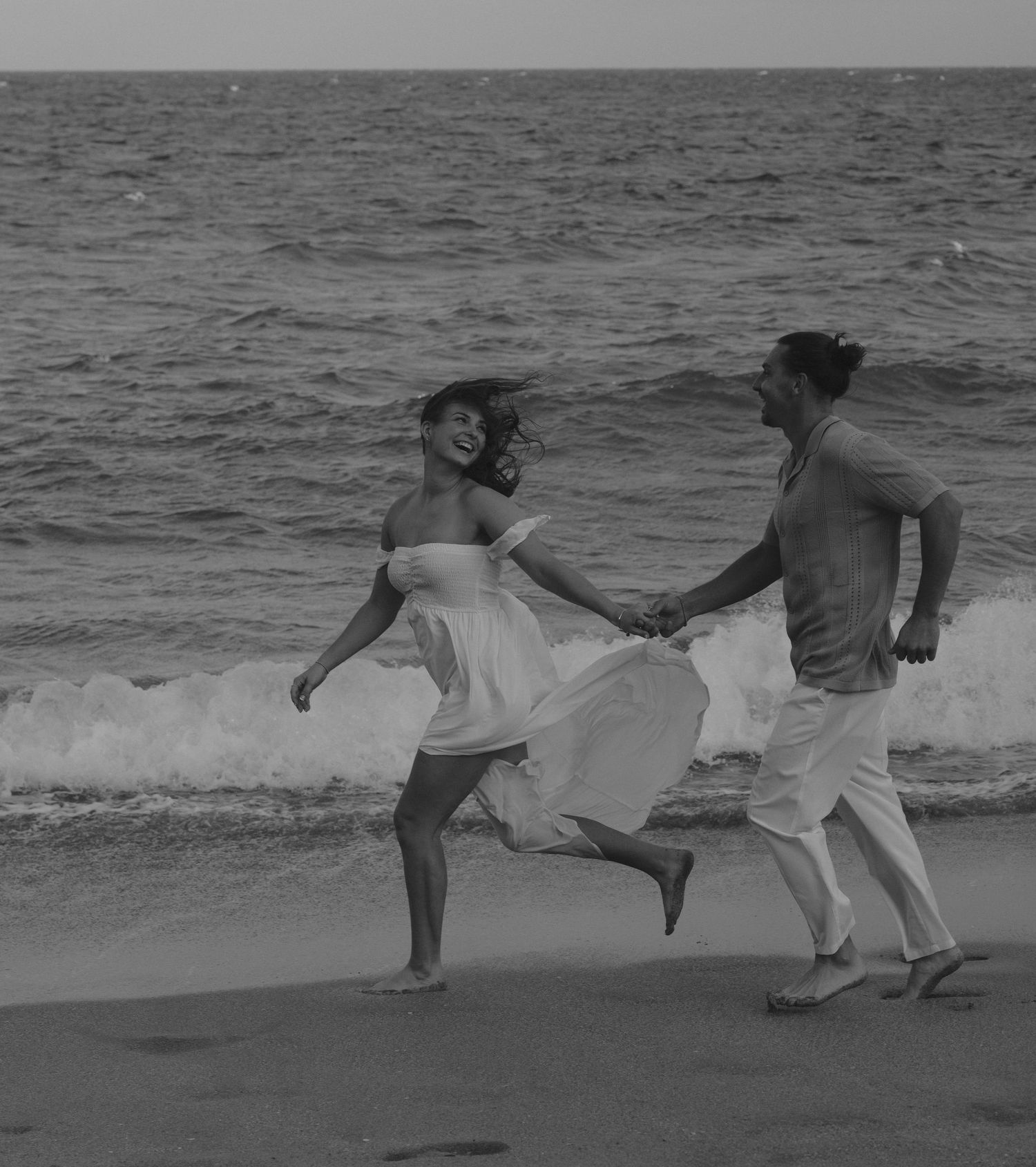 A black and white image of a couple joyfully running along a beach shore with waves in the background.
