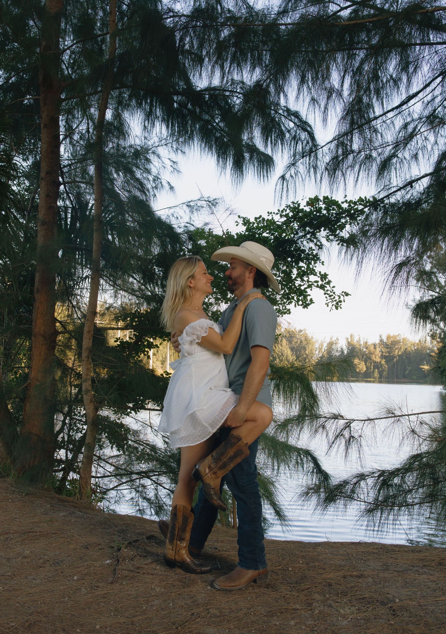 A romantic couple poses by a lakeside at sunset, surrounded by pine trees with the water glistening behind them.
