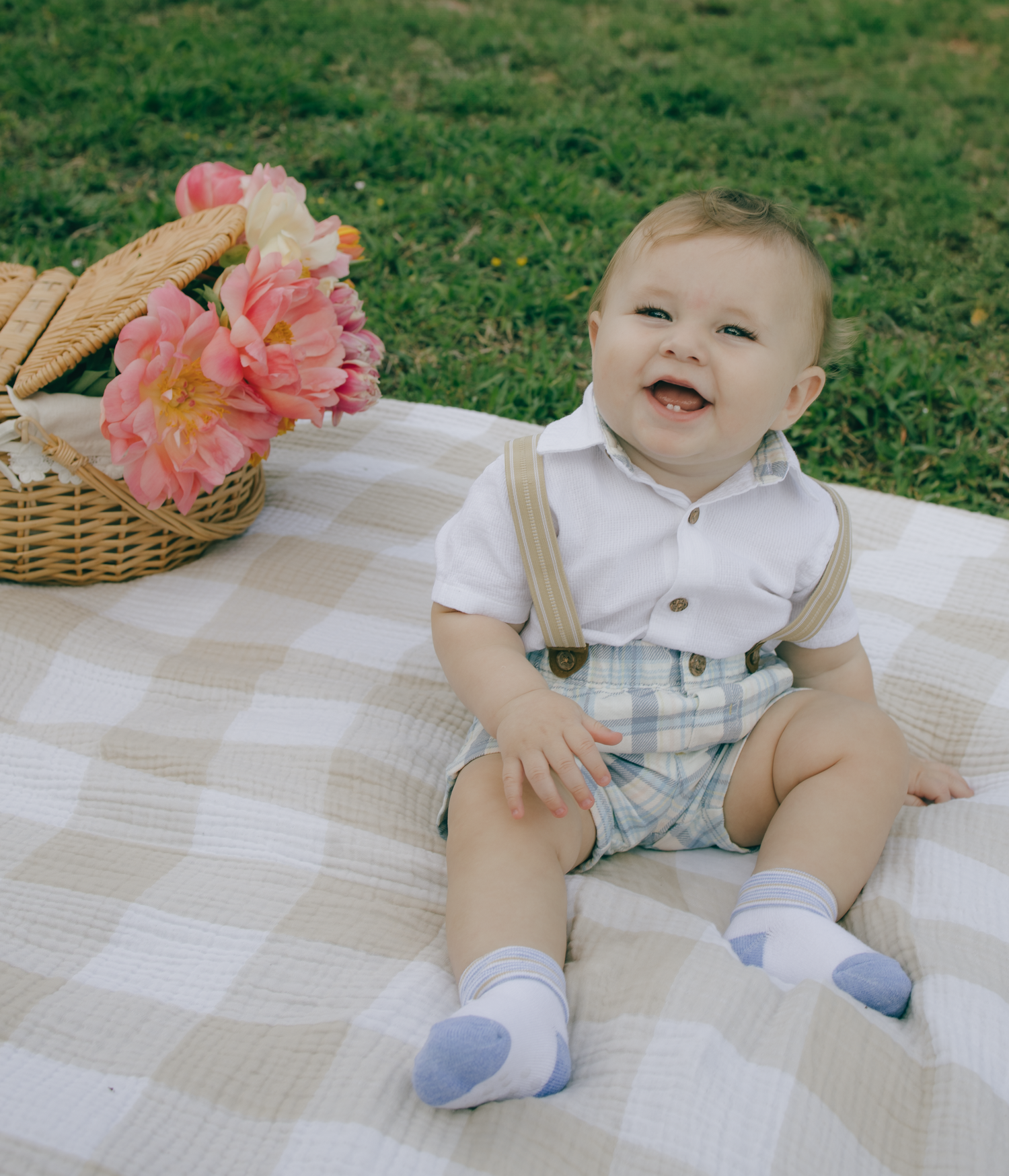 A smiling baby in suspenders sits on a checkered blanket next to pink flowers in a basket.