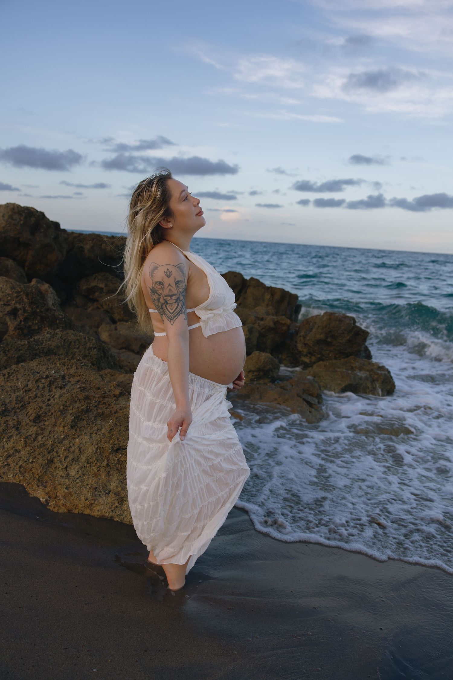 Expectant mother in white dress poses on rocky beach at sunset with ocean waves crashing behind.