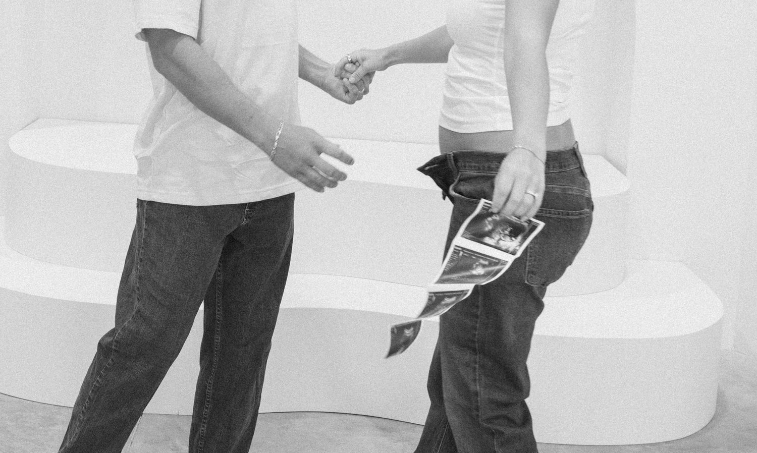 Black and white photo of a couple holding hands, woman holding ultrasound photos, expecting parents announcement.