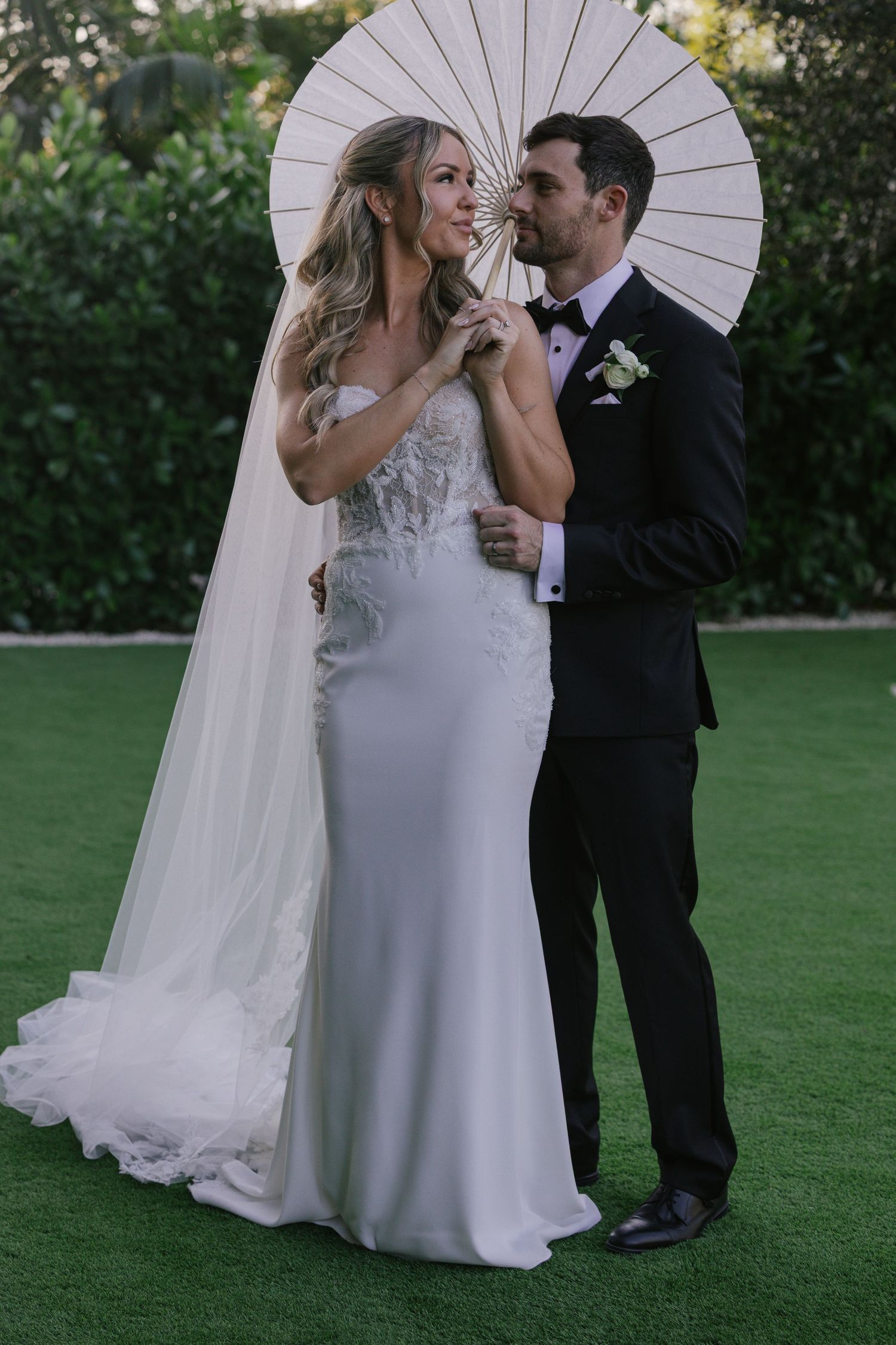 Wedding couple shares intimate moments under white parasol umbrellas in a lush garden setting.