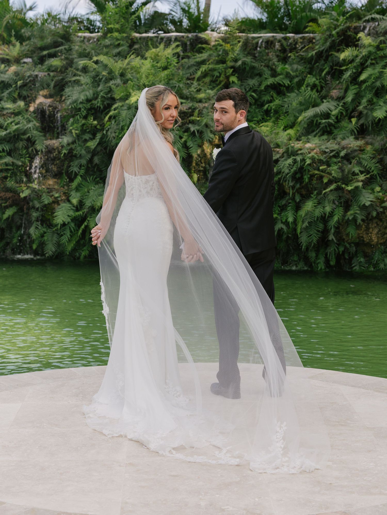 A couple in wedding attire walks along a scenic garden path with a tranquil green pond in the background.