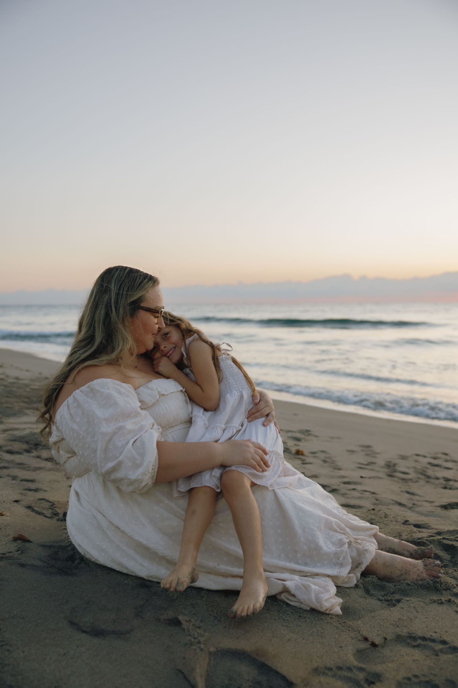 Two people in white outfits share a tender moment while sitting on the sandy beach at dusk.