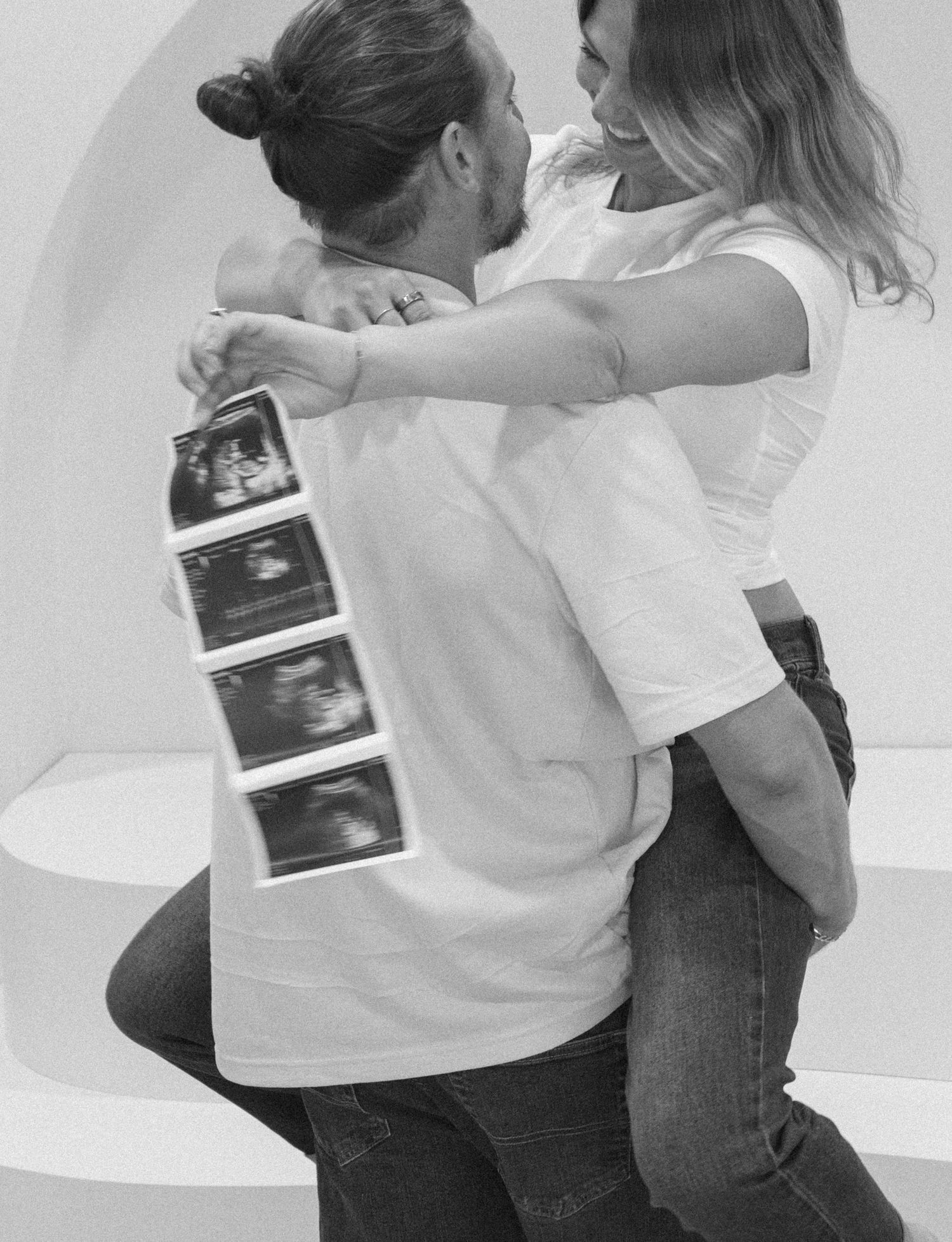 Black and white photo of a couple embracing joyfully while holding ultrasound scan photos.