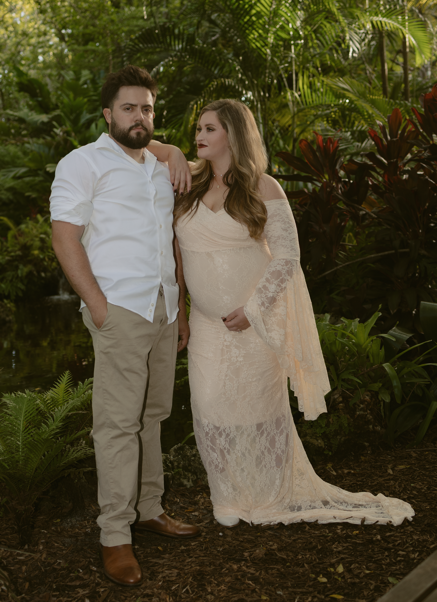 A couple in white attire pose together in a lush garden setting with natural lighting and tropical foliage.