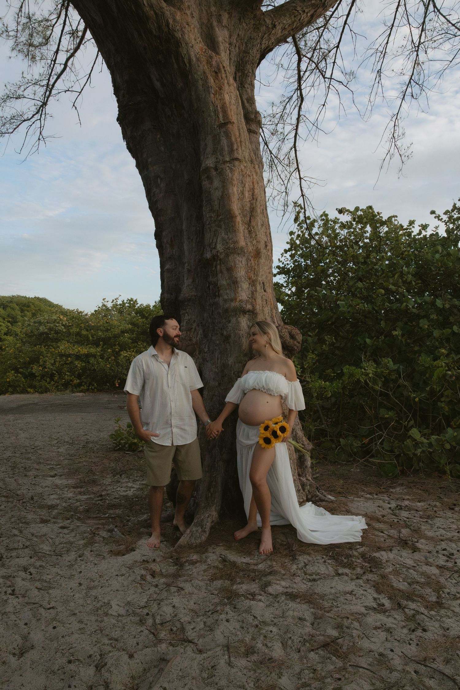 Romantic couple poses against large tree trunk in natural outdoor setting with sunflowers and flowing fabric.
