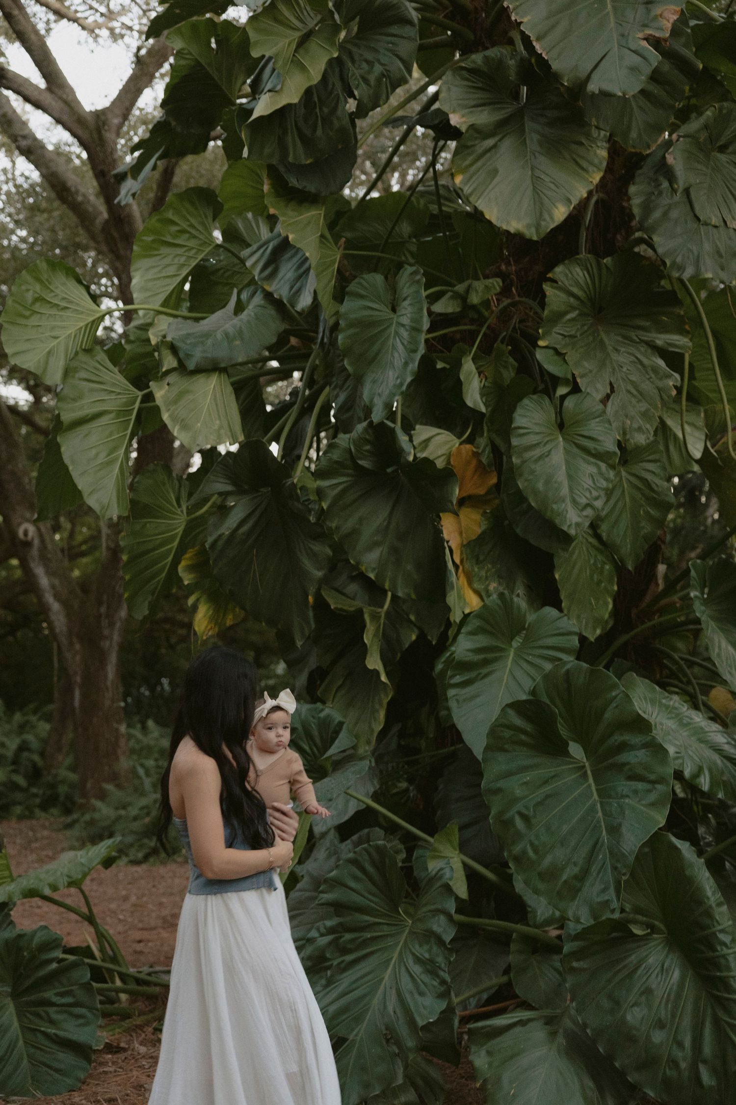 A woman in a white dress stands among large tropical monstera leaves in a garden setting.