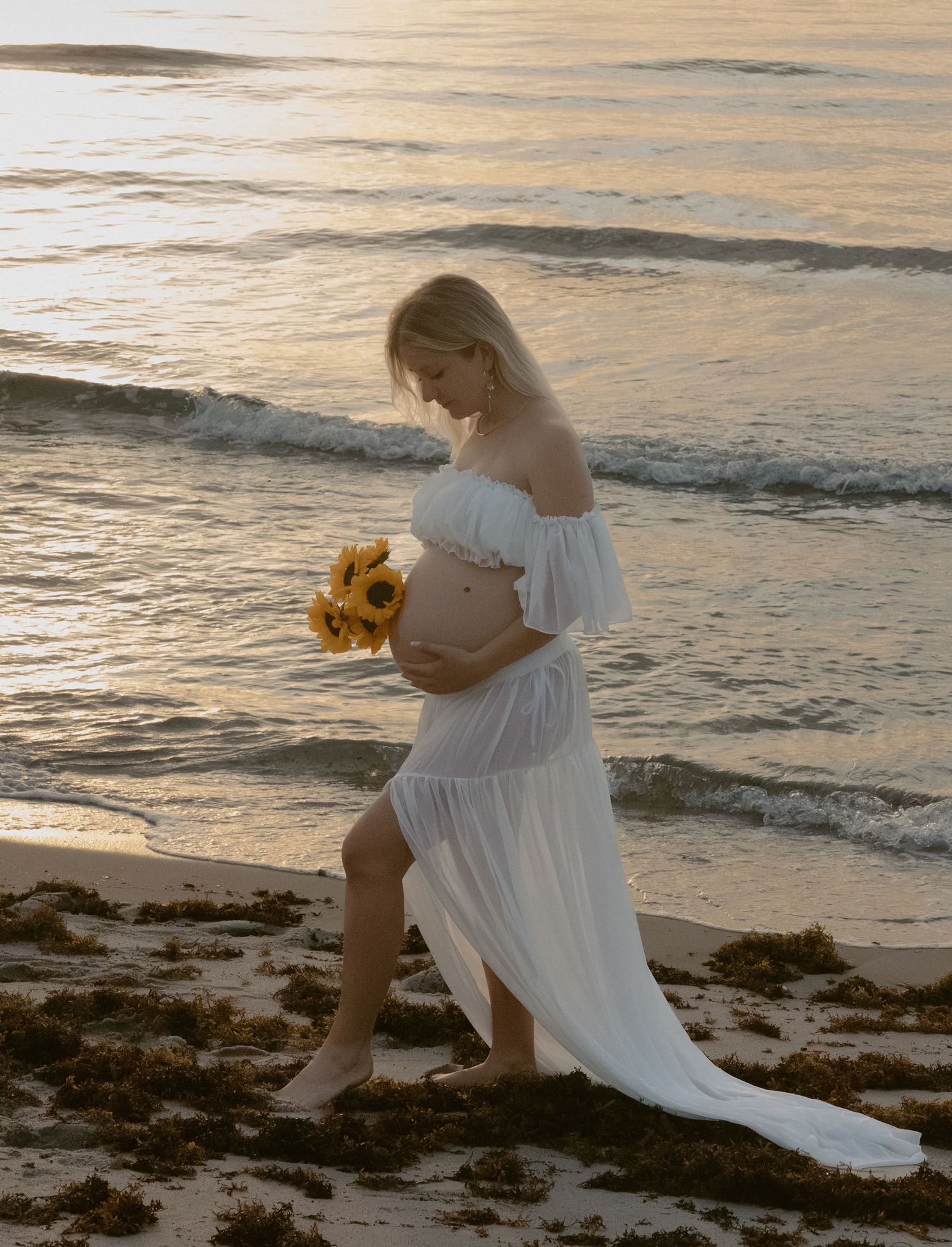 A pregnant woman in a flowing white dress holds sunflowers while standing on a beach at sunset.