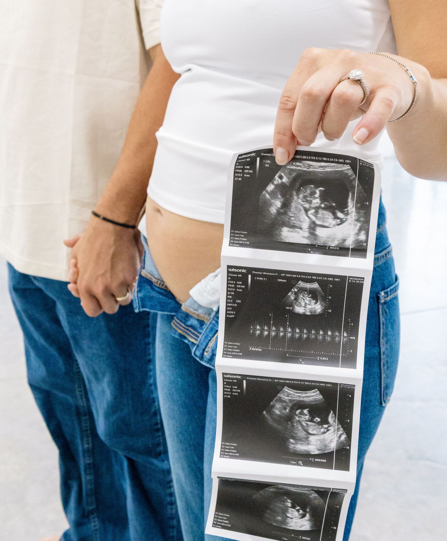 Pregnant woman holding ultrasound photos while standing with partner, both wearing jeans and white tops.