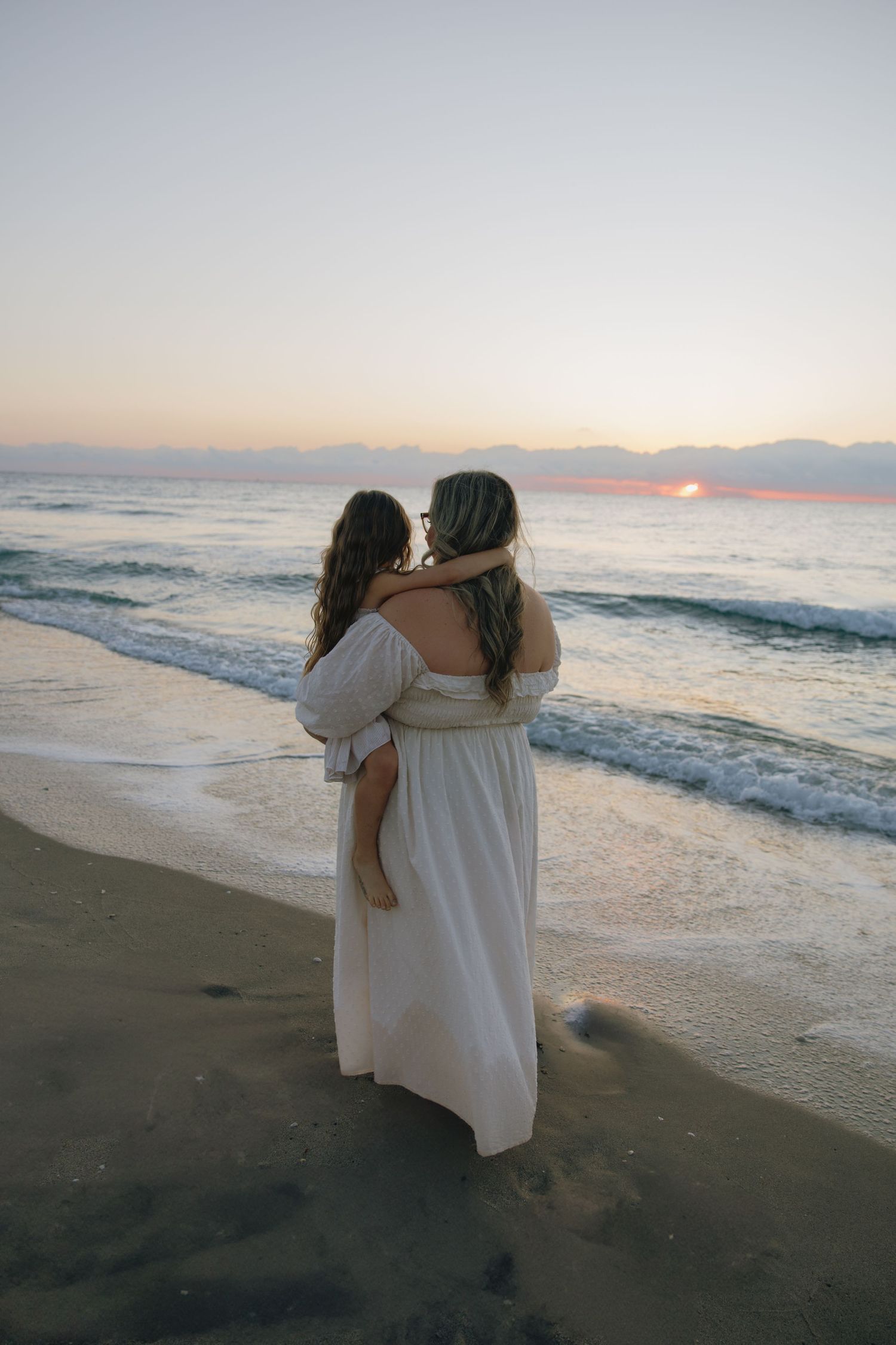 Two people in white dresses embrace while watching the sunset over ocean waves.