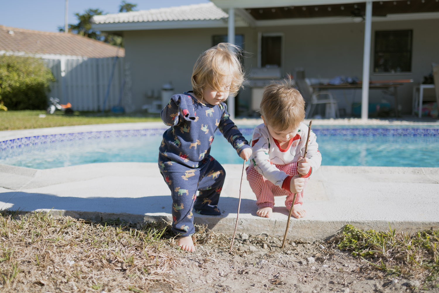 Two young children in overalls playing together near a backyard swimming pool.