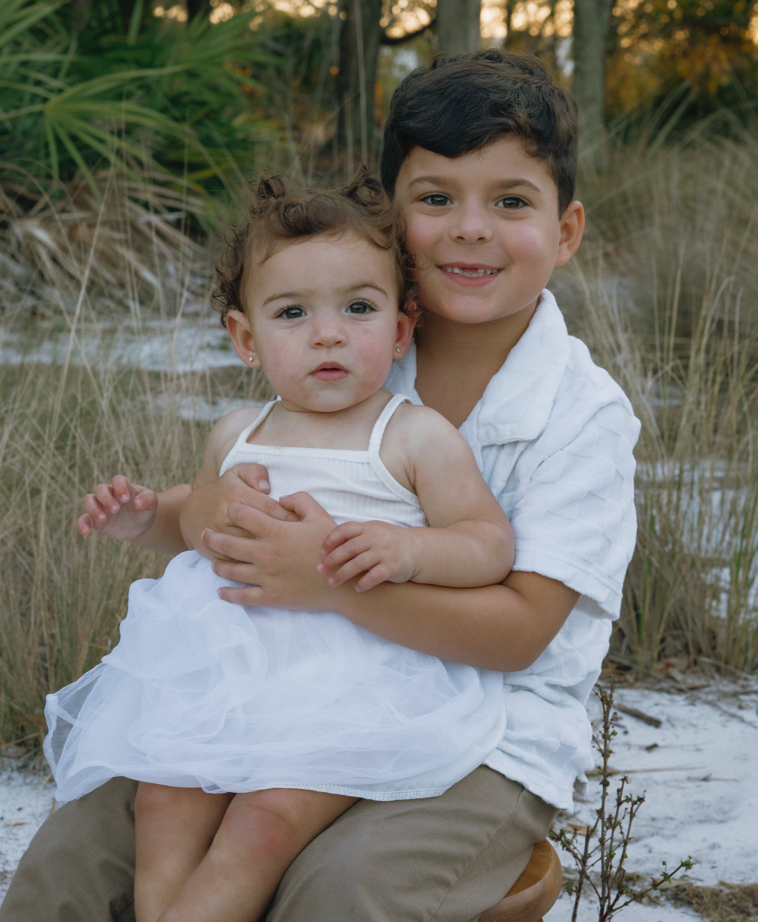 Two children in white clothing sitting together on a sandy beach at sunset.