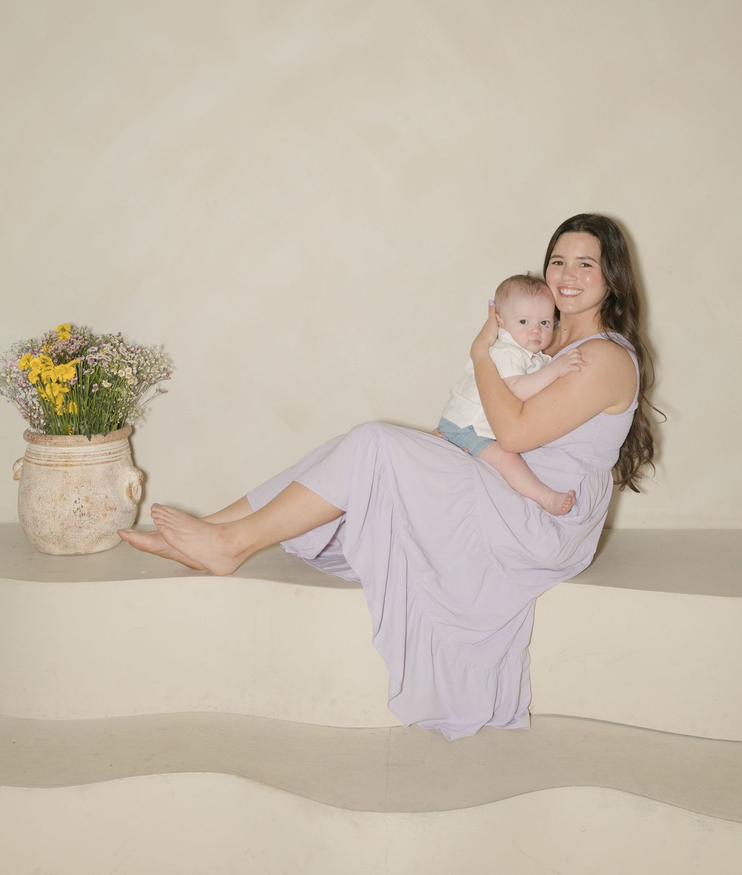 Woman in lavender dress holding baby, seated beside rustic vase with yellow flowers in soft neutral setting.