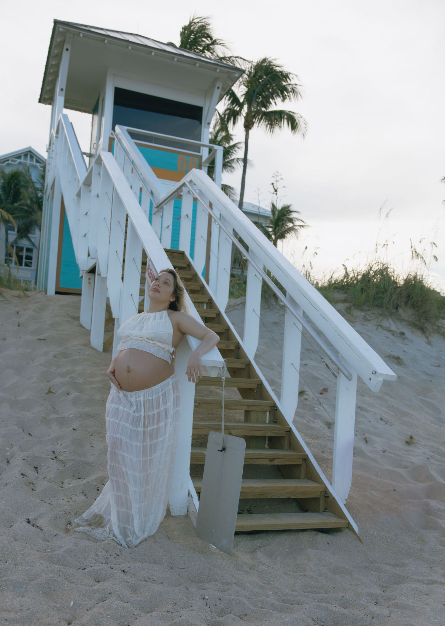 White-dressed figure dances by coastal stairway with turquoise building in background.