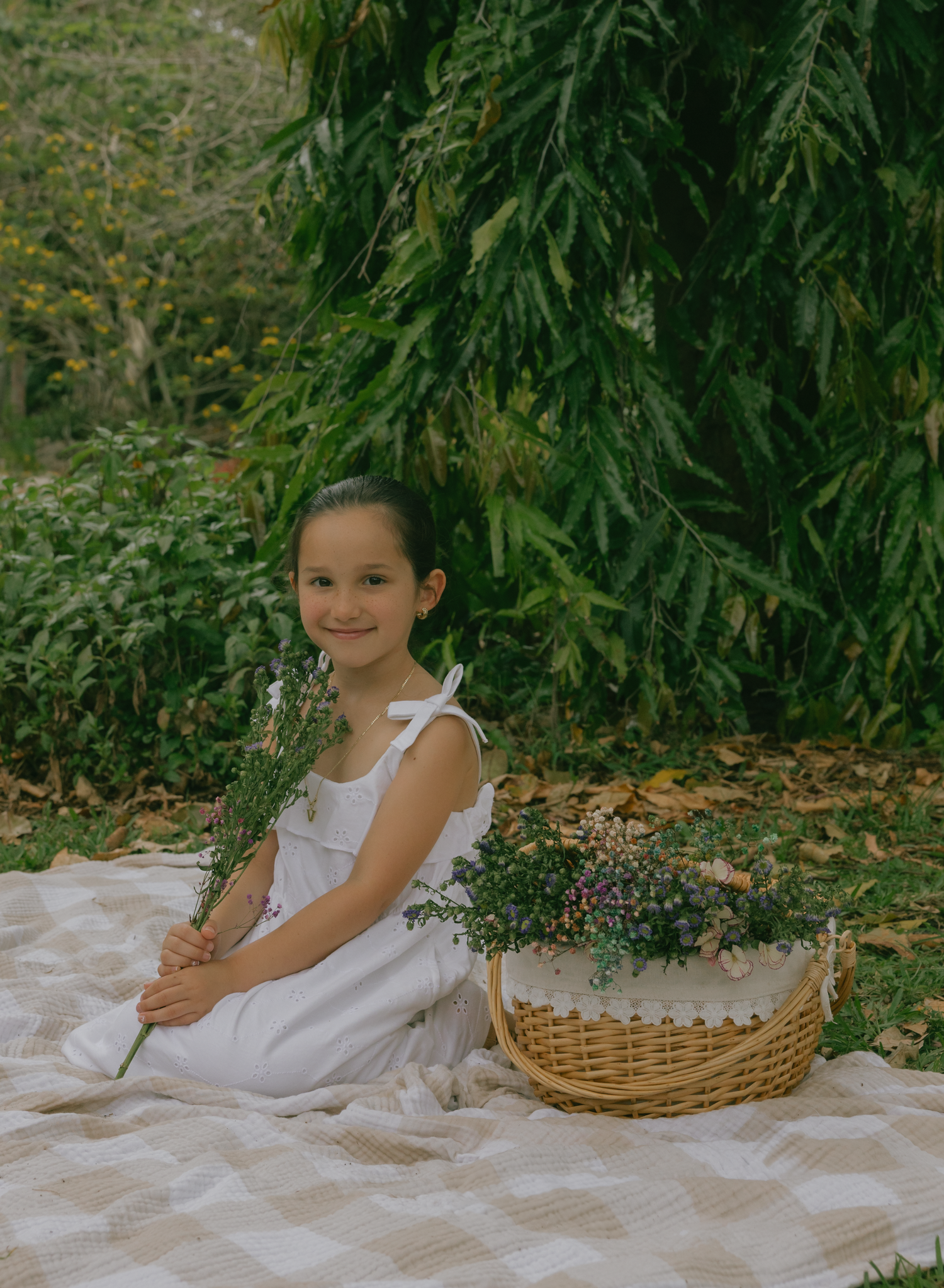 Smiling girl in white dress sitting with flowers beside picnic basket under willow tree.