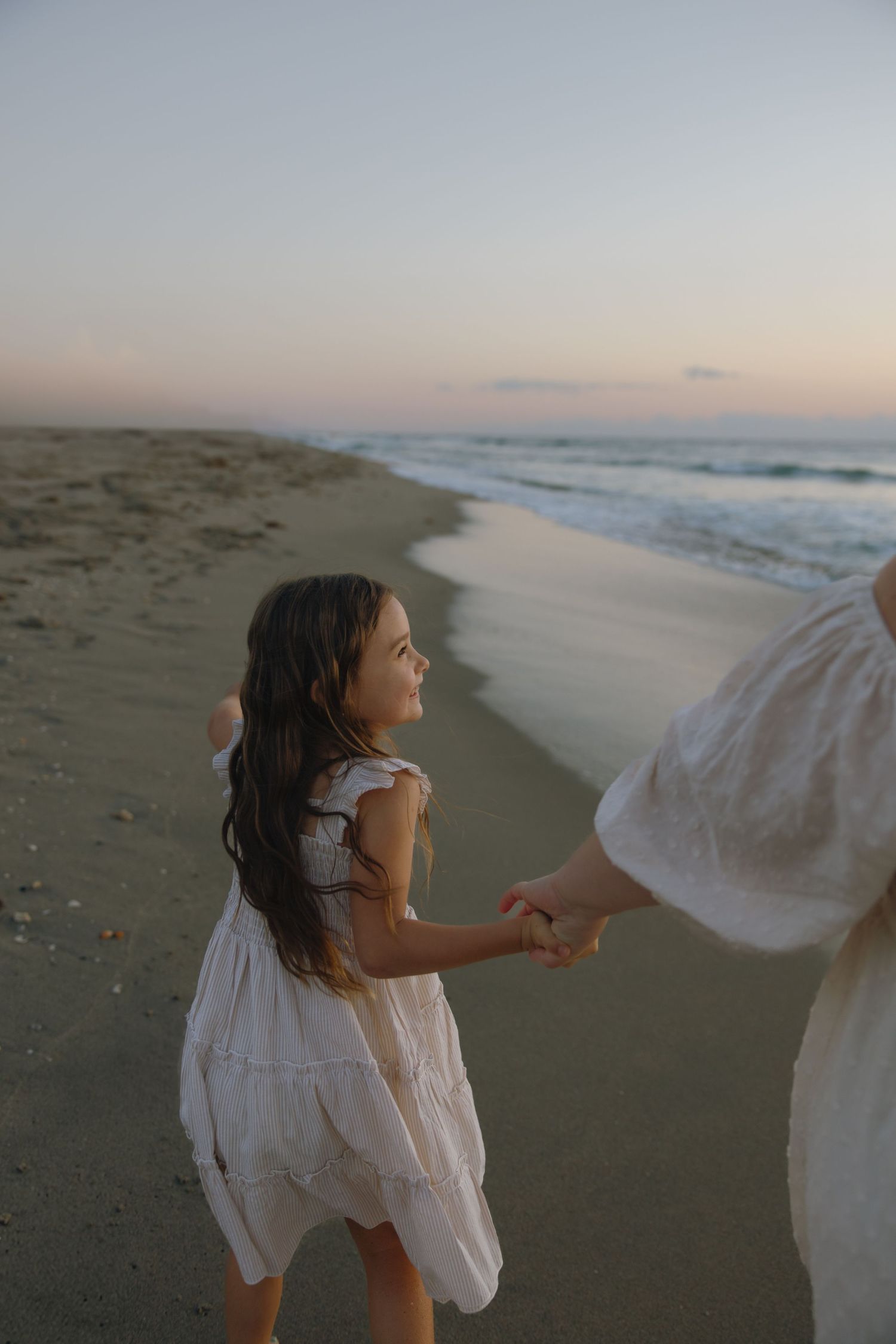 A figure in a flowing white dress holds hands with someone while walking along a peaceful beach at sunset.