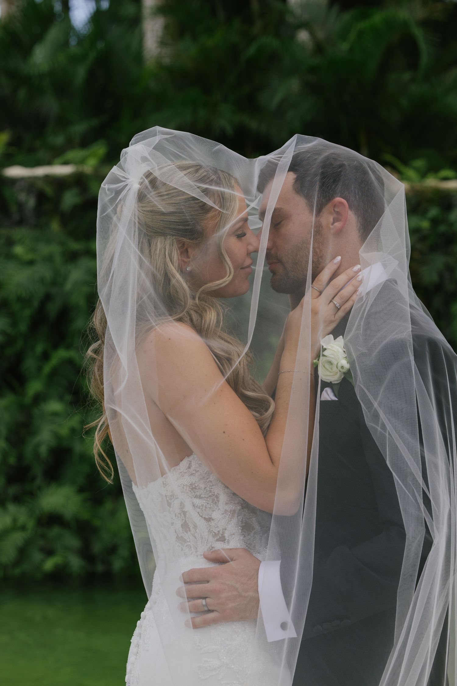 A romantic wedding couple shares a tender kiss under a flowing white veil in an outdoor garden setting.