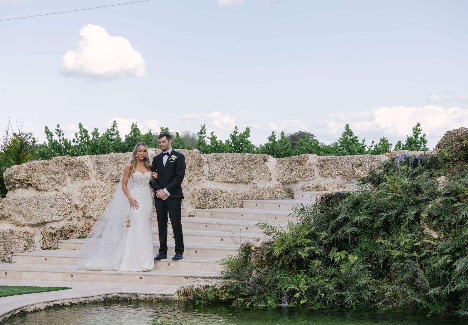 A bride and groom stand together on stone steps surrounded by lush greenery at an outdoor wedding venue.