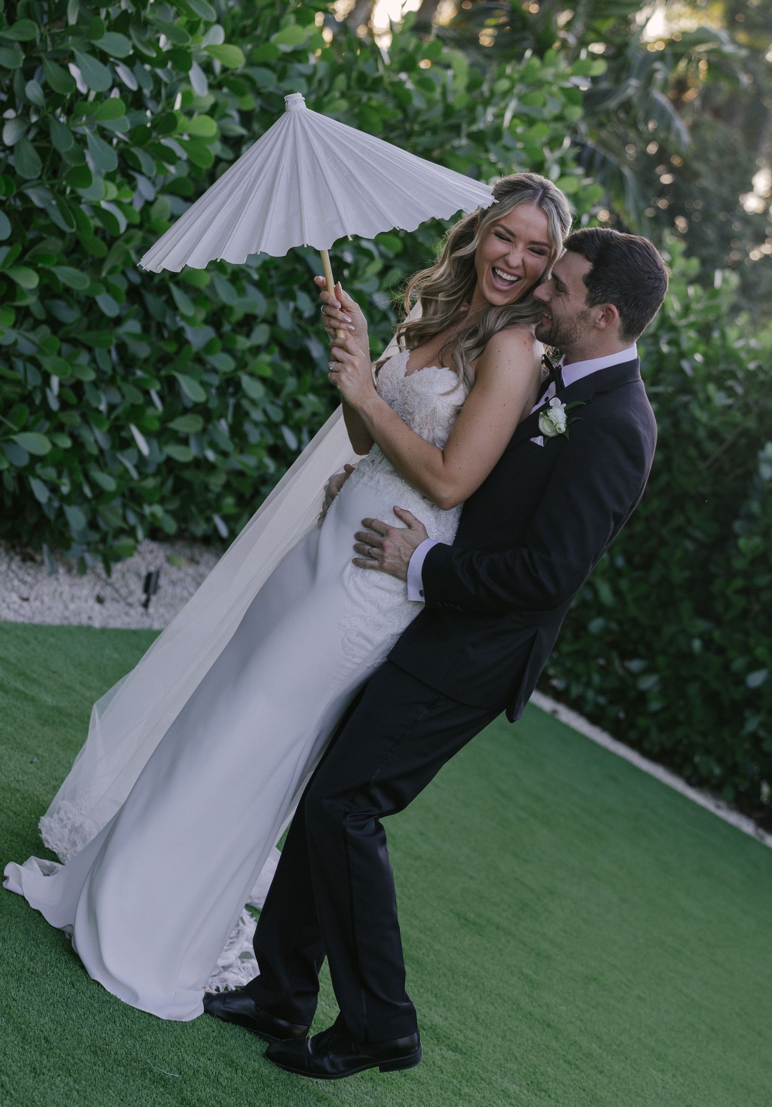 A romantic wedding couple dances together in a garden setting with a white parasol.