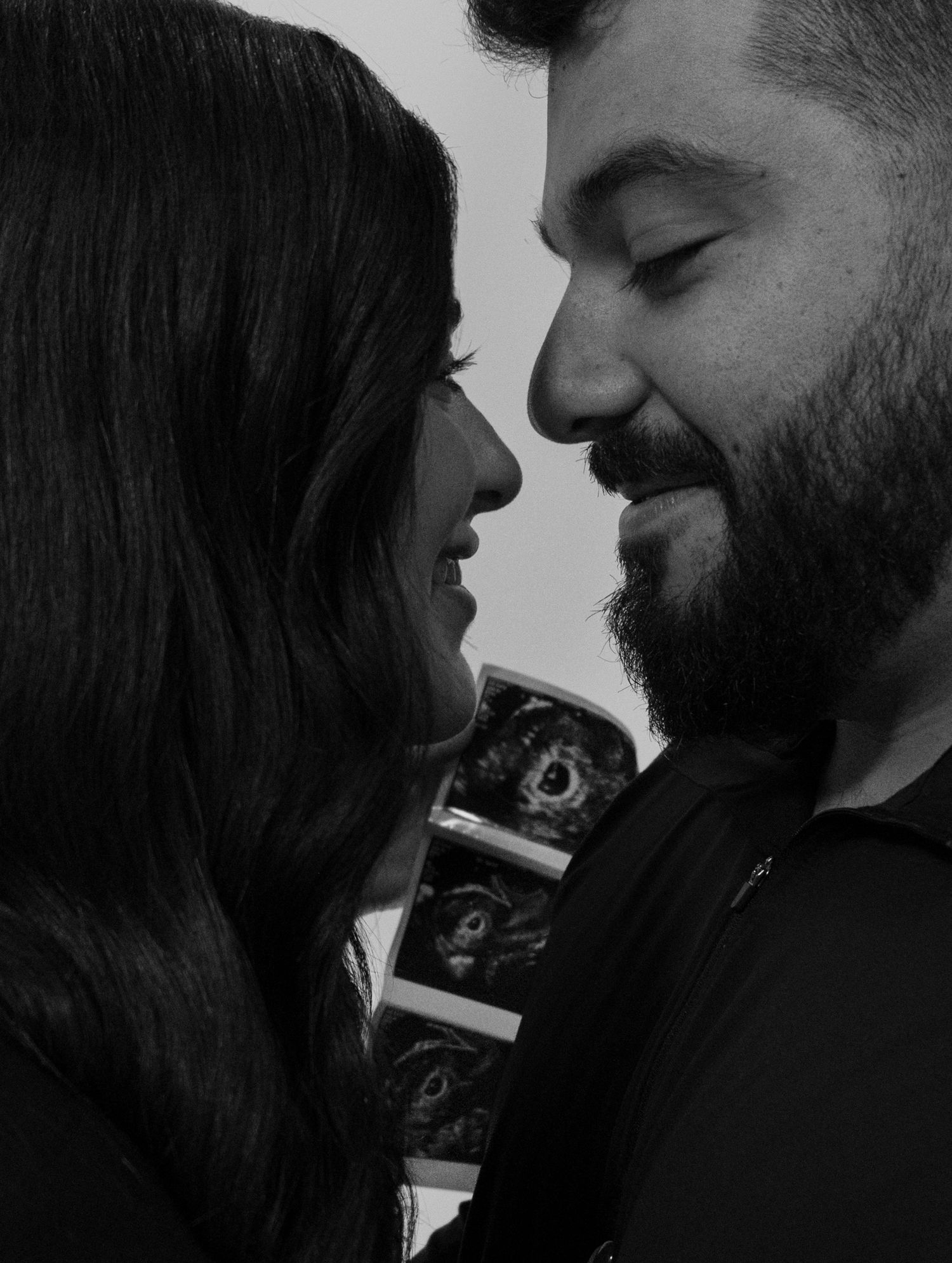 Black and white photo of a smiling couple facing each other with ultrasound photos visible in the background.