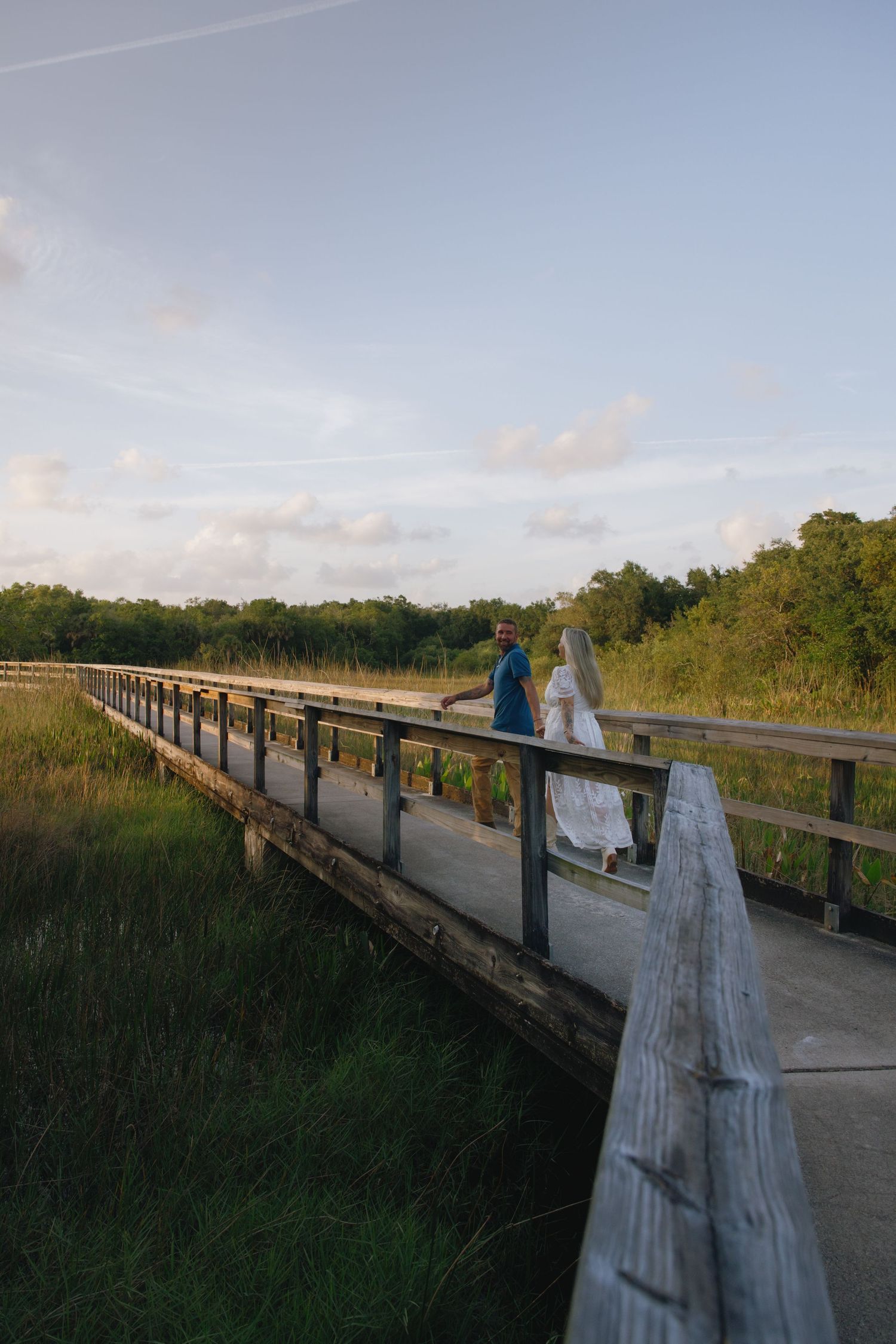 A couple walks along a wooden boardwalk during sunset in a natural coastal setting.