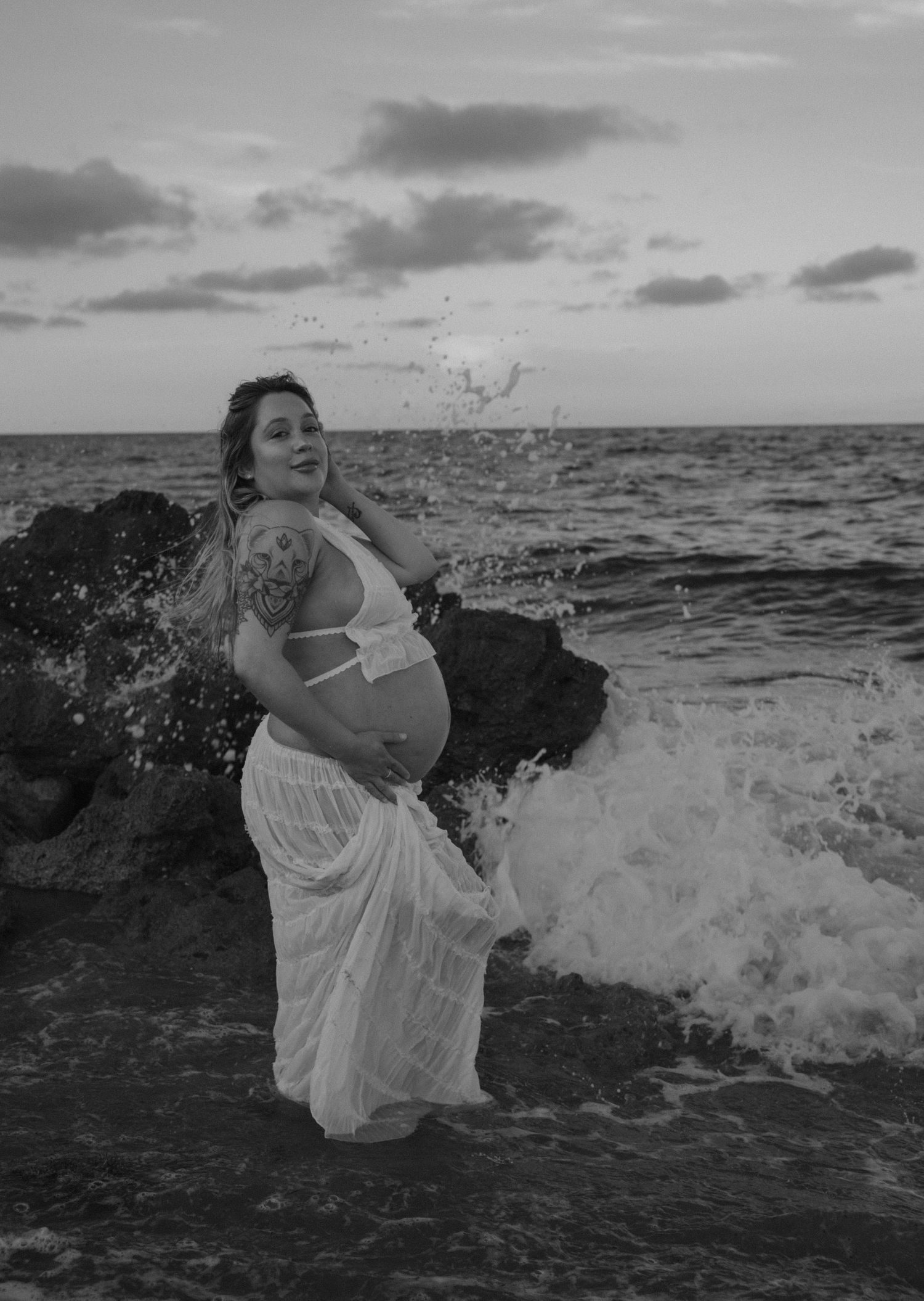 Artistic black and white maternity photo with ocean waves splashing against rocky shore.