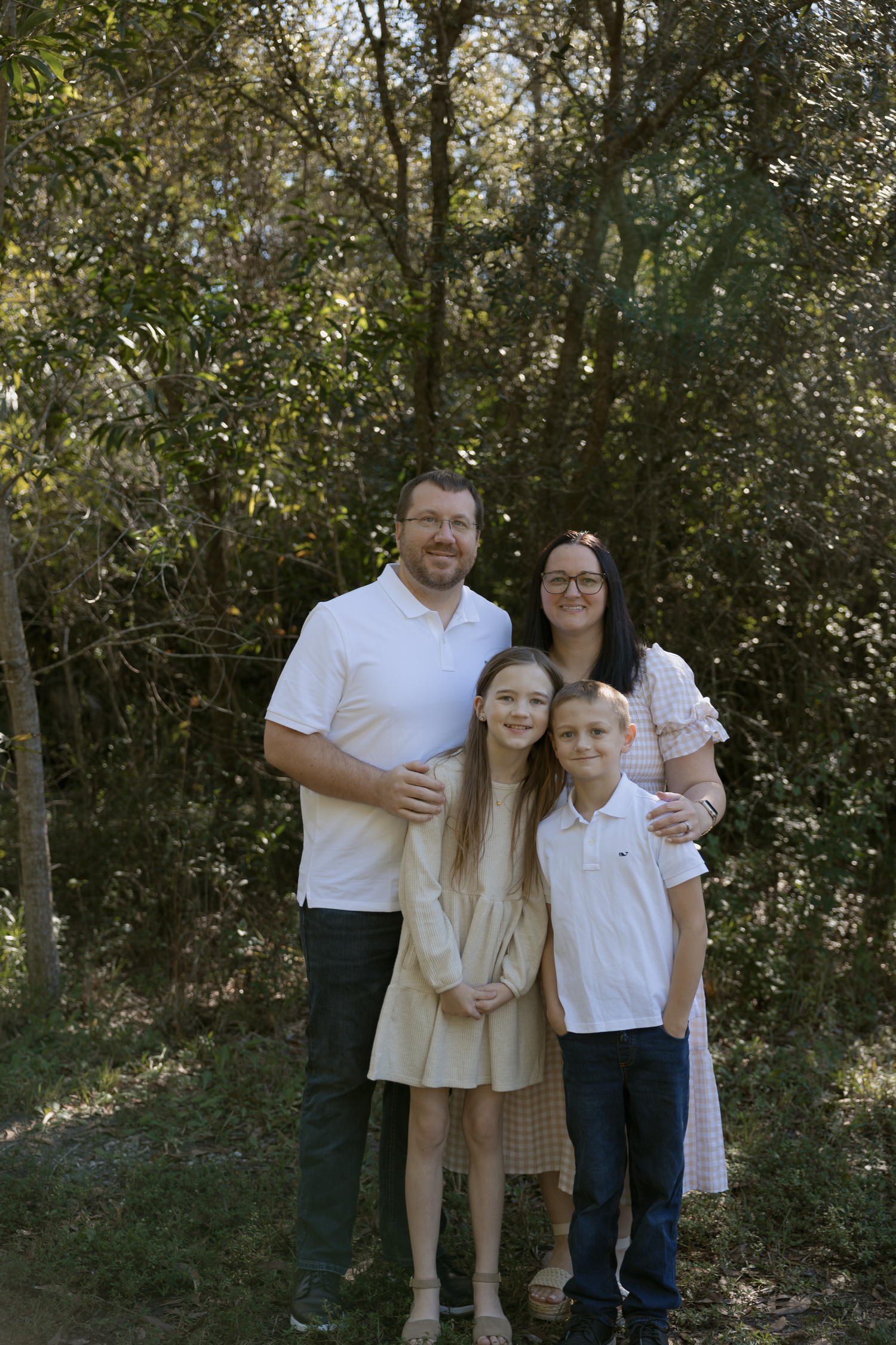 A family in coordinated white and beige outfits poses together in front of trees during an outdoor photo session.