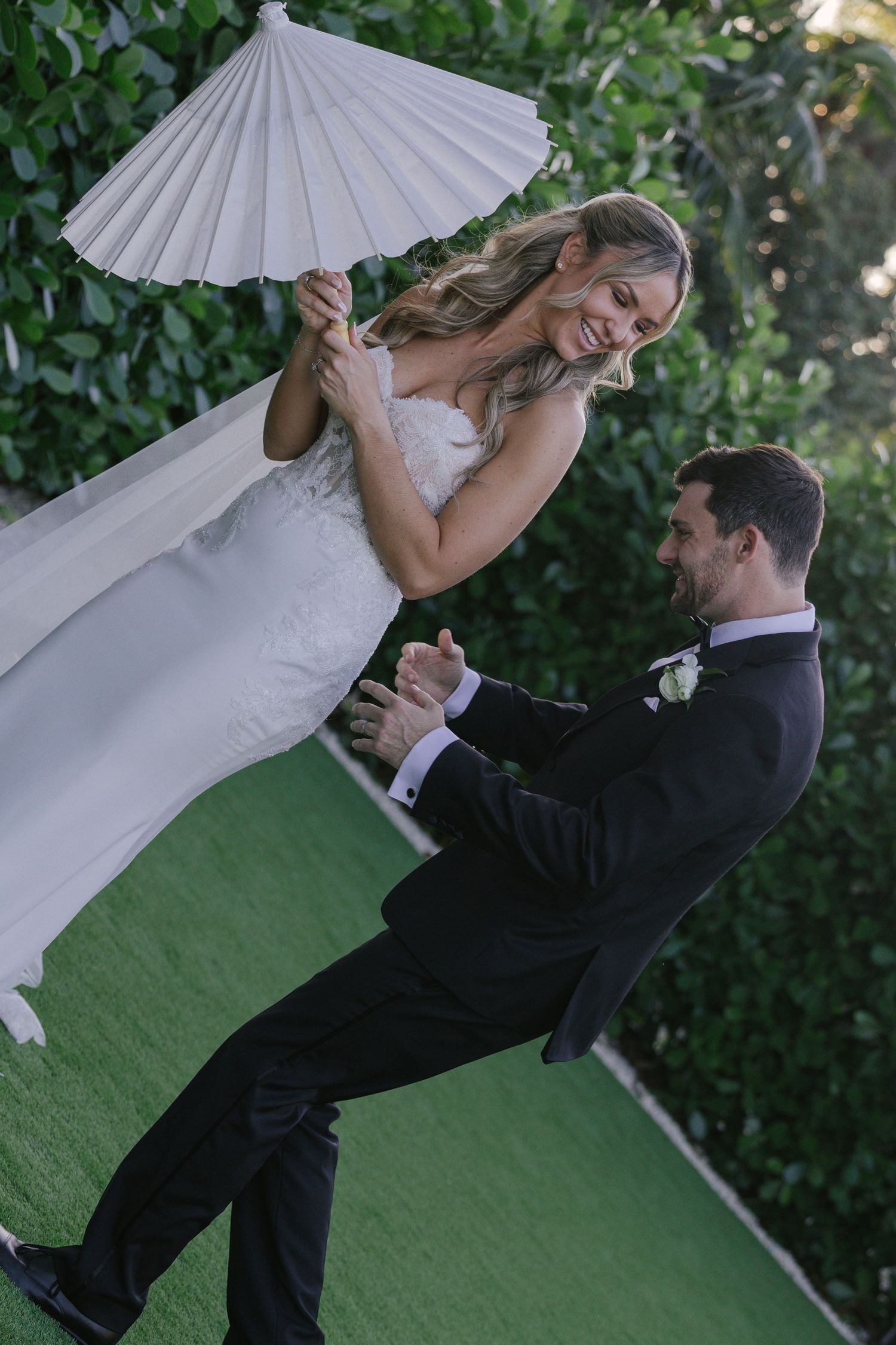 A couple shares a romantic moment under a white paper parasol in an outdoor garden setting.