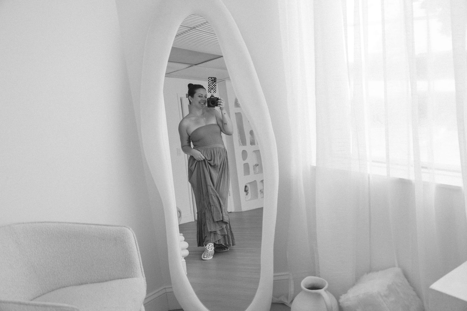Black and white photo of a woman taking a selfie in a large ornate mirror in a bright, modern room.