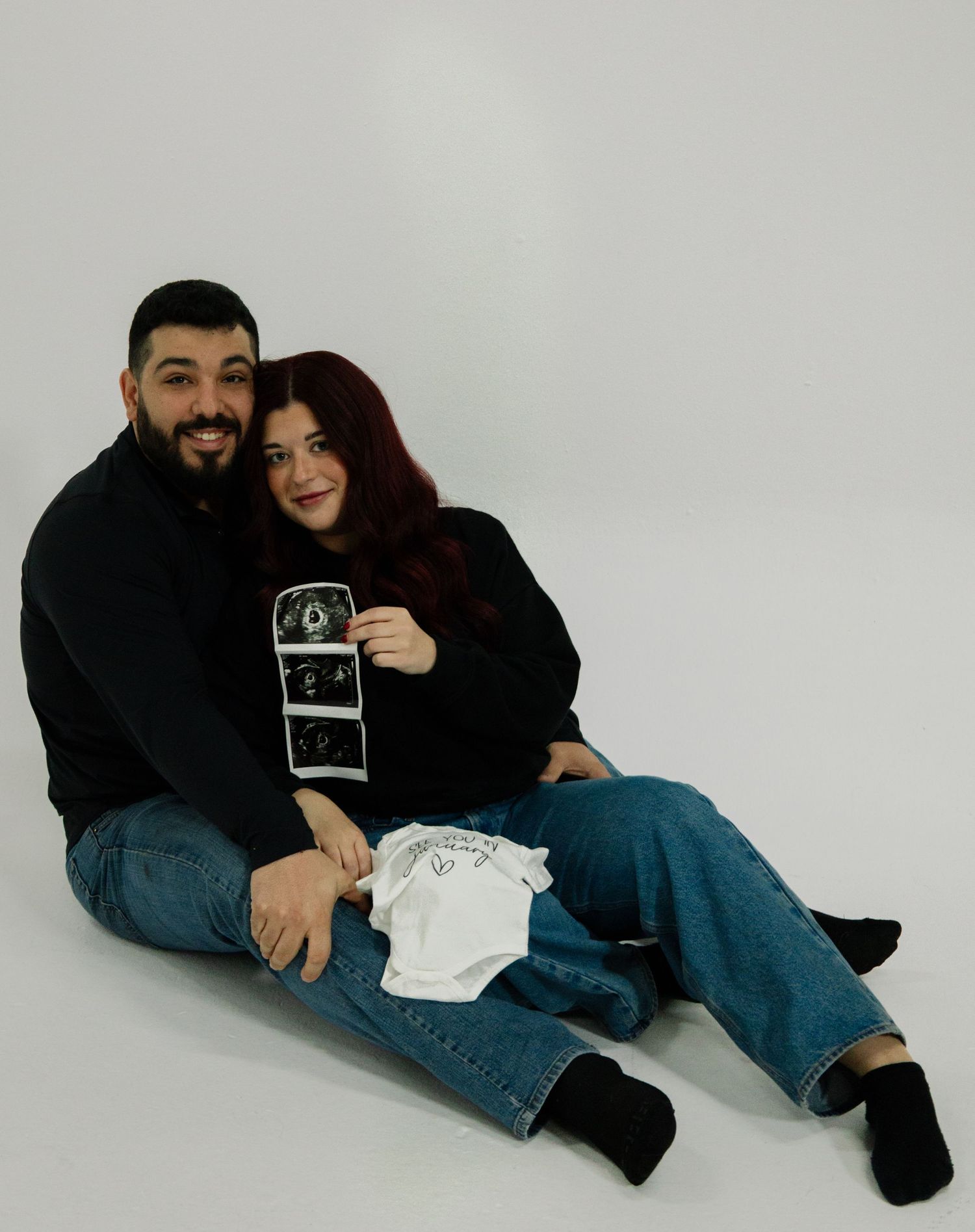 Couple sitting on floor holding baby ultrasound photos and white onesie, announcing pregnancy in matching black outfits.