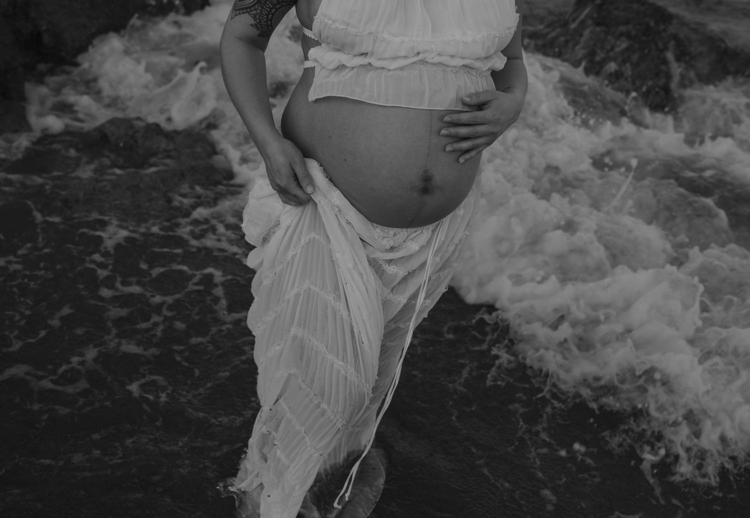 Black and white close-up of pregnant belly with waves crashing in background.