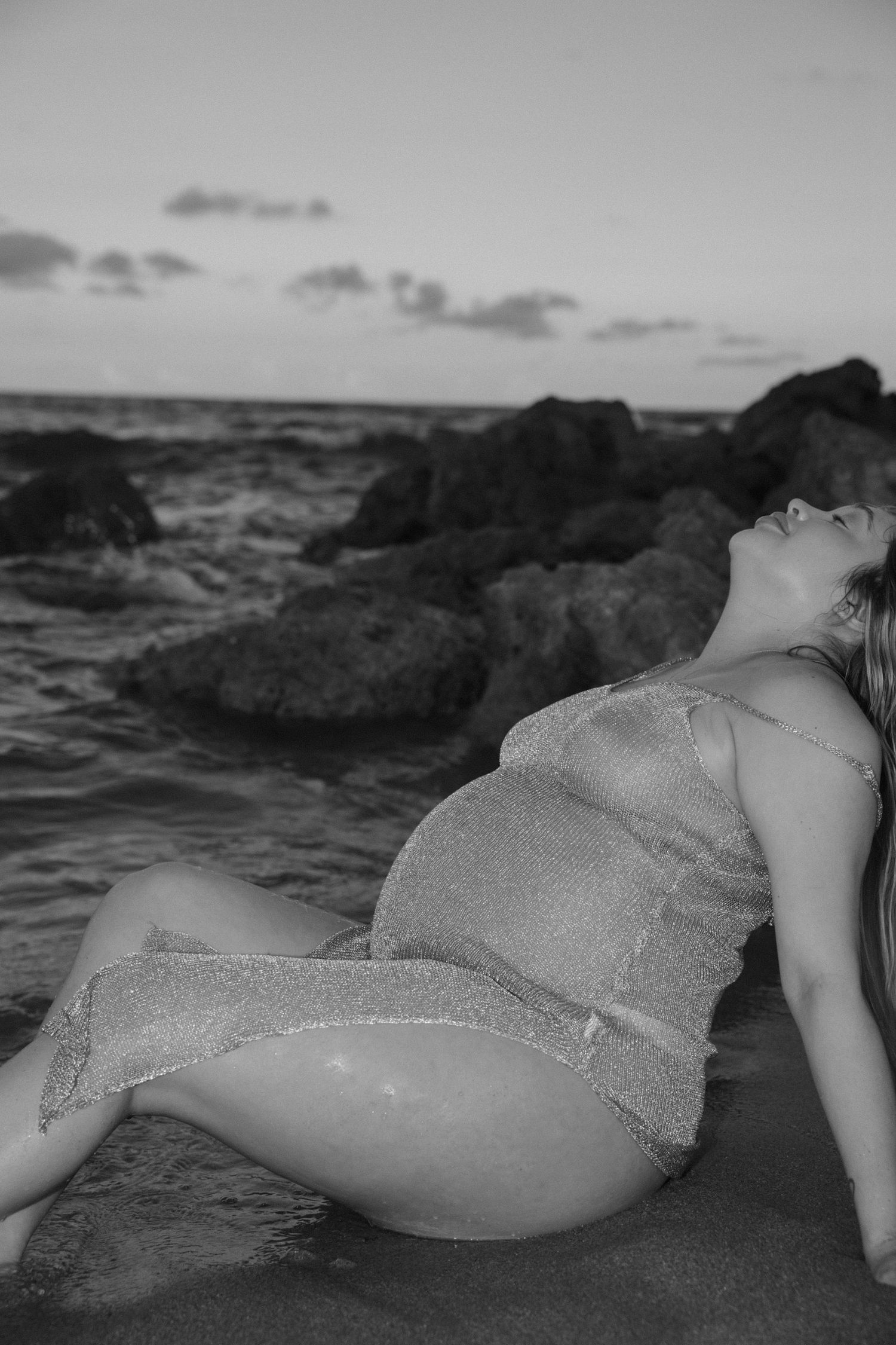 Dramatic black and white maternity portrait with ocean waves and rocky coastline.