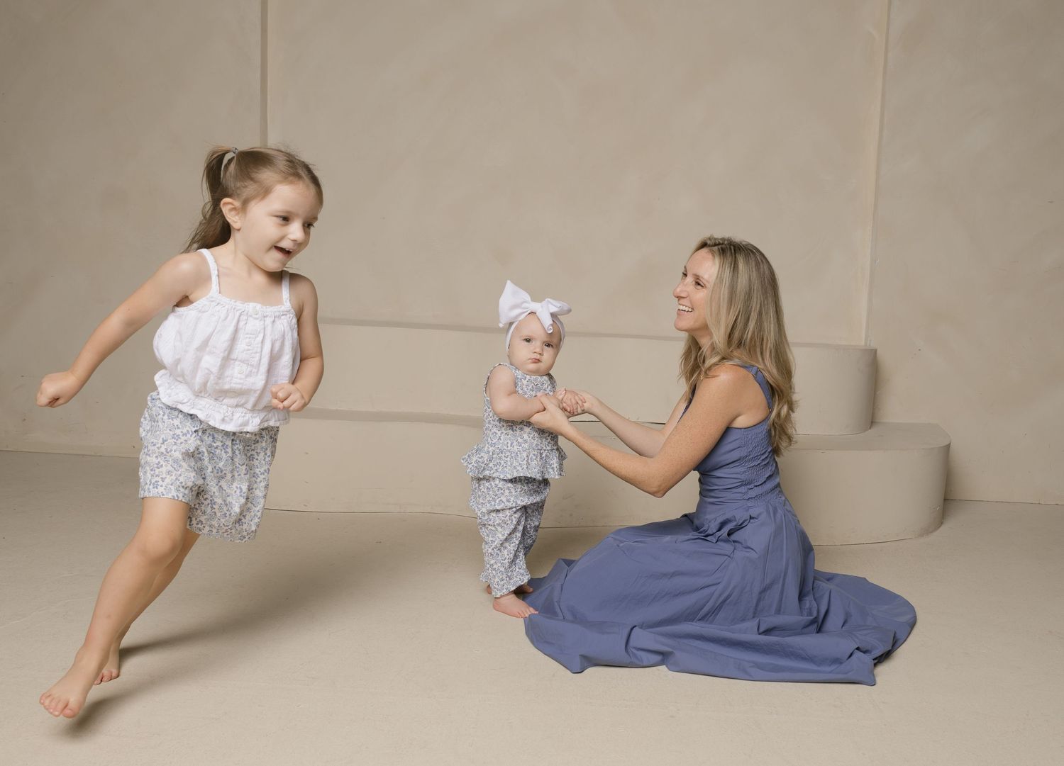 A mother sits on the floor smiling as her toddler runs toward her and baby in matching gray outfits.