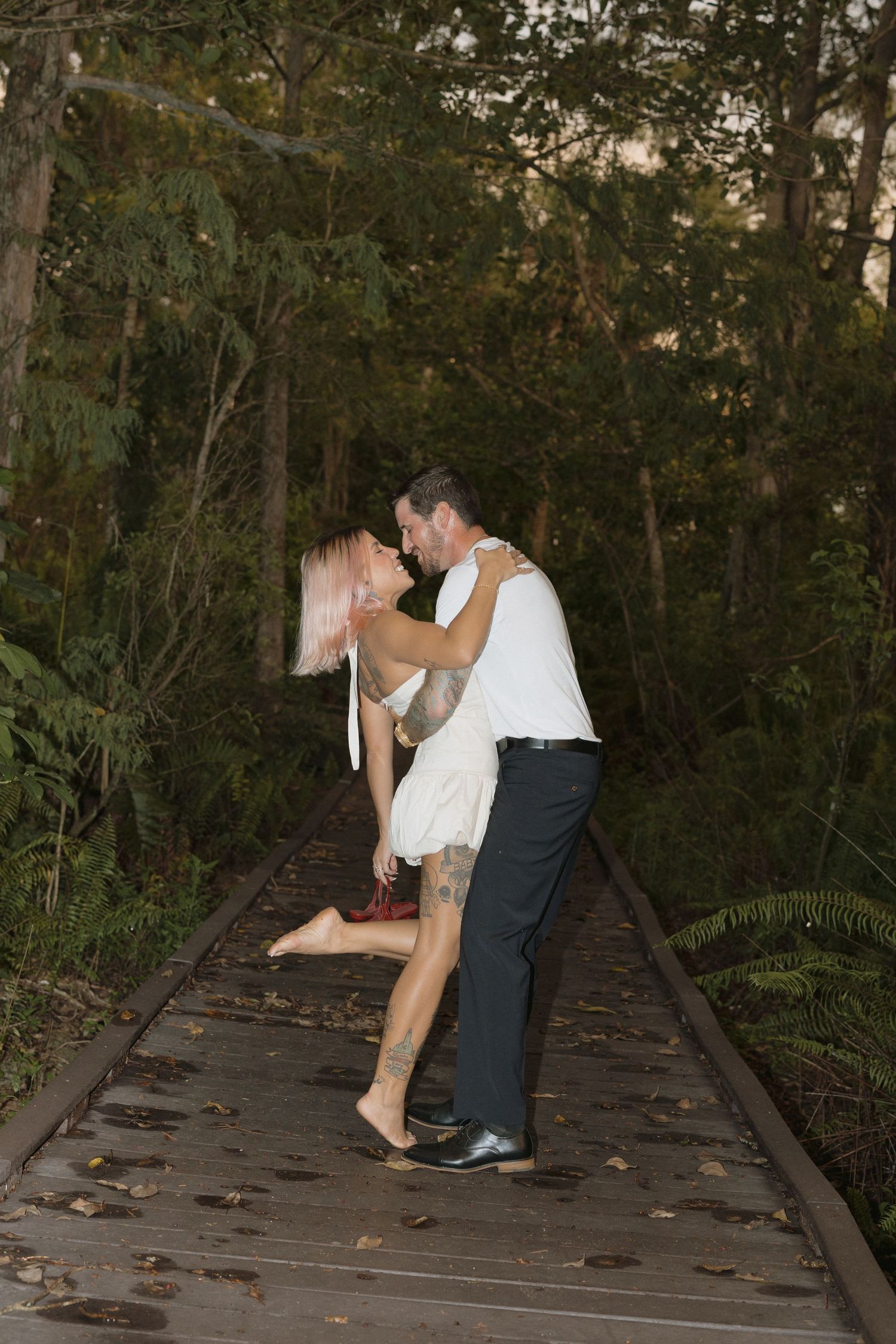 A romantic couple shares a dip kiss pose on a wooden boardwalk surrounded by lush forest greenery.