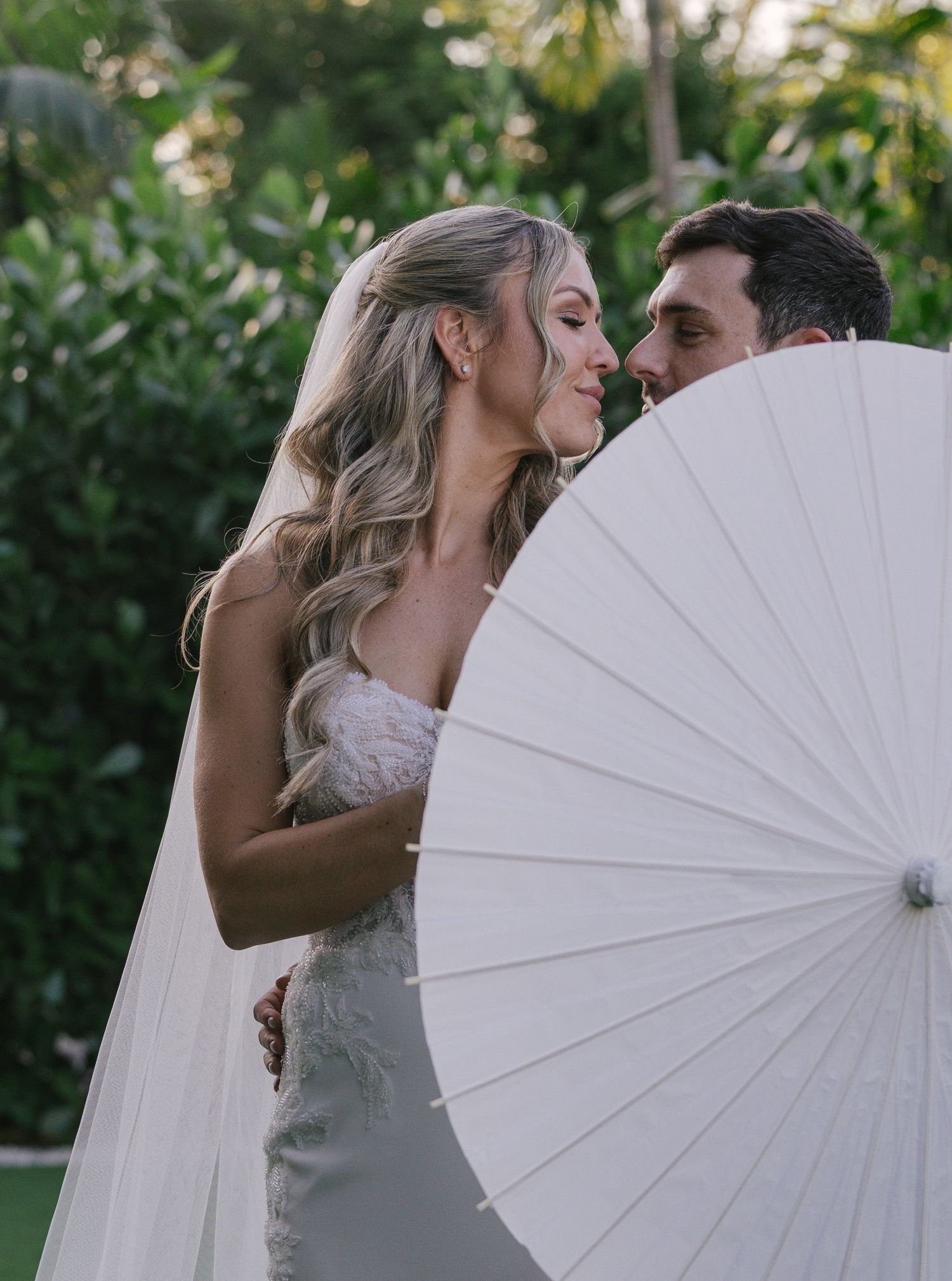 A couple shares a romantic kiss behind a white parasol in a garden setting during their wedding photoshoot.