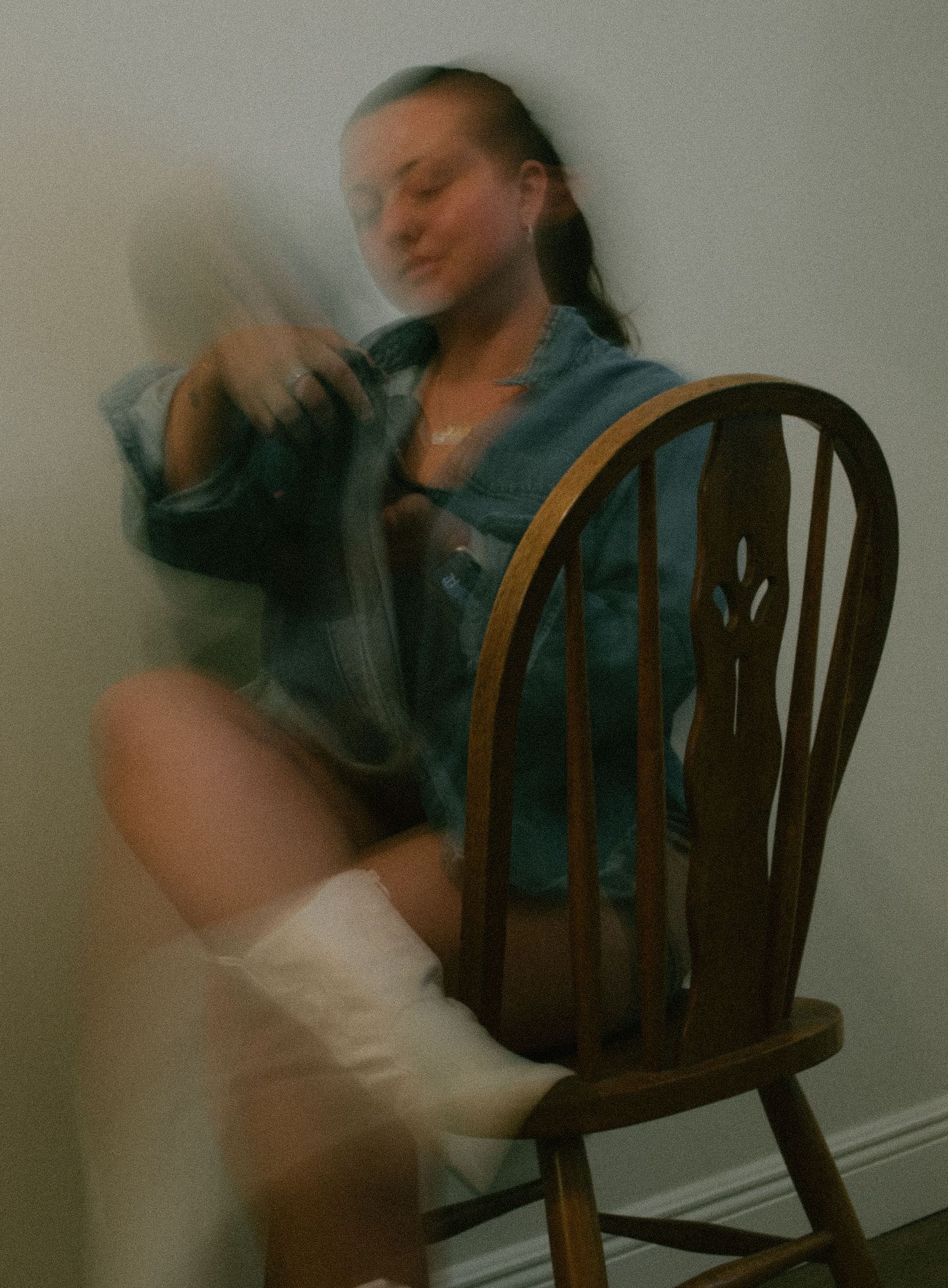 A person in a denim jacket sits on a wooden Windsor chair wearing tall white boots in a softly blurred vintage style photo.