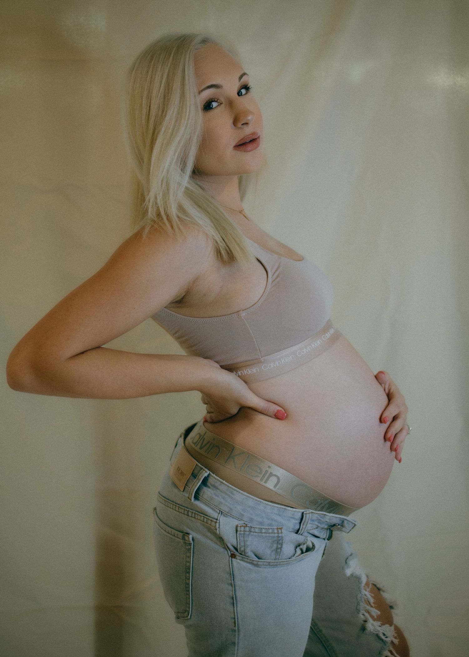 Maternity photo showing pregnant woman in casual clothing against warm backdrop.