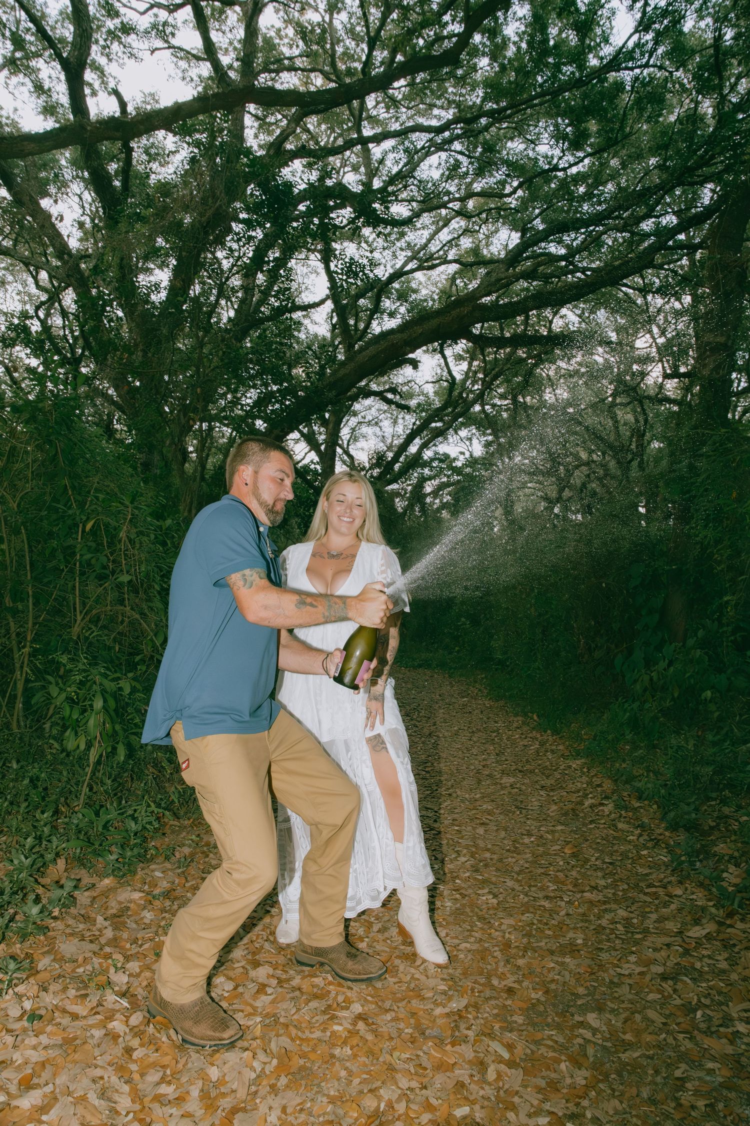 Couple celebrating with champagne on a tree-lined path.