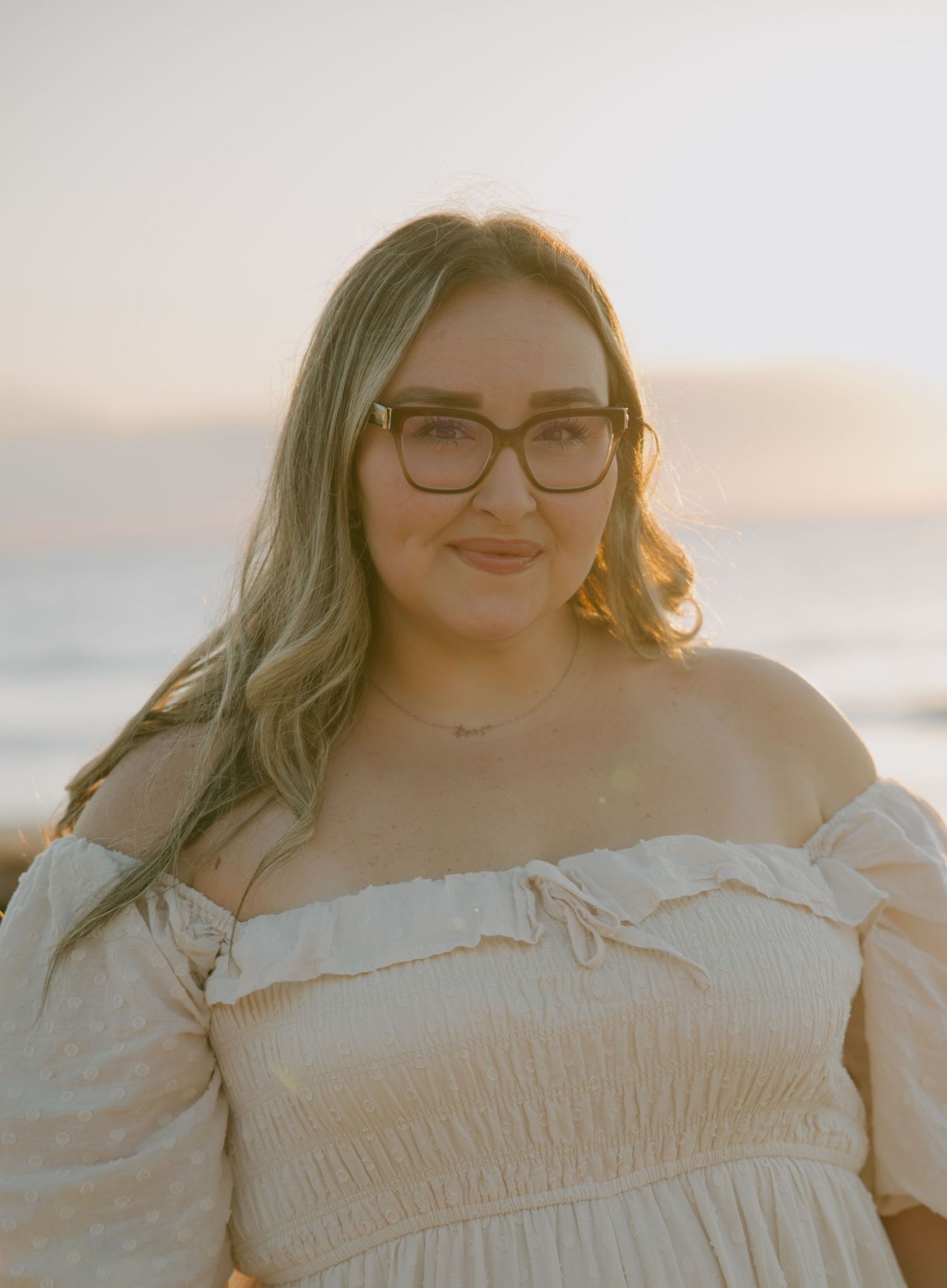 A person in a white off-shoulder dress smiles warmly at the beach during sunset.