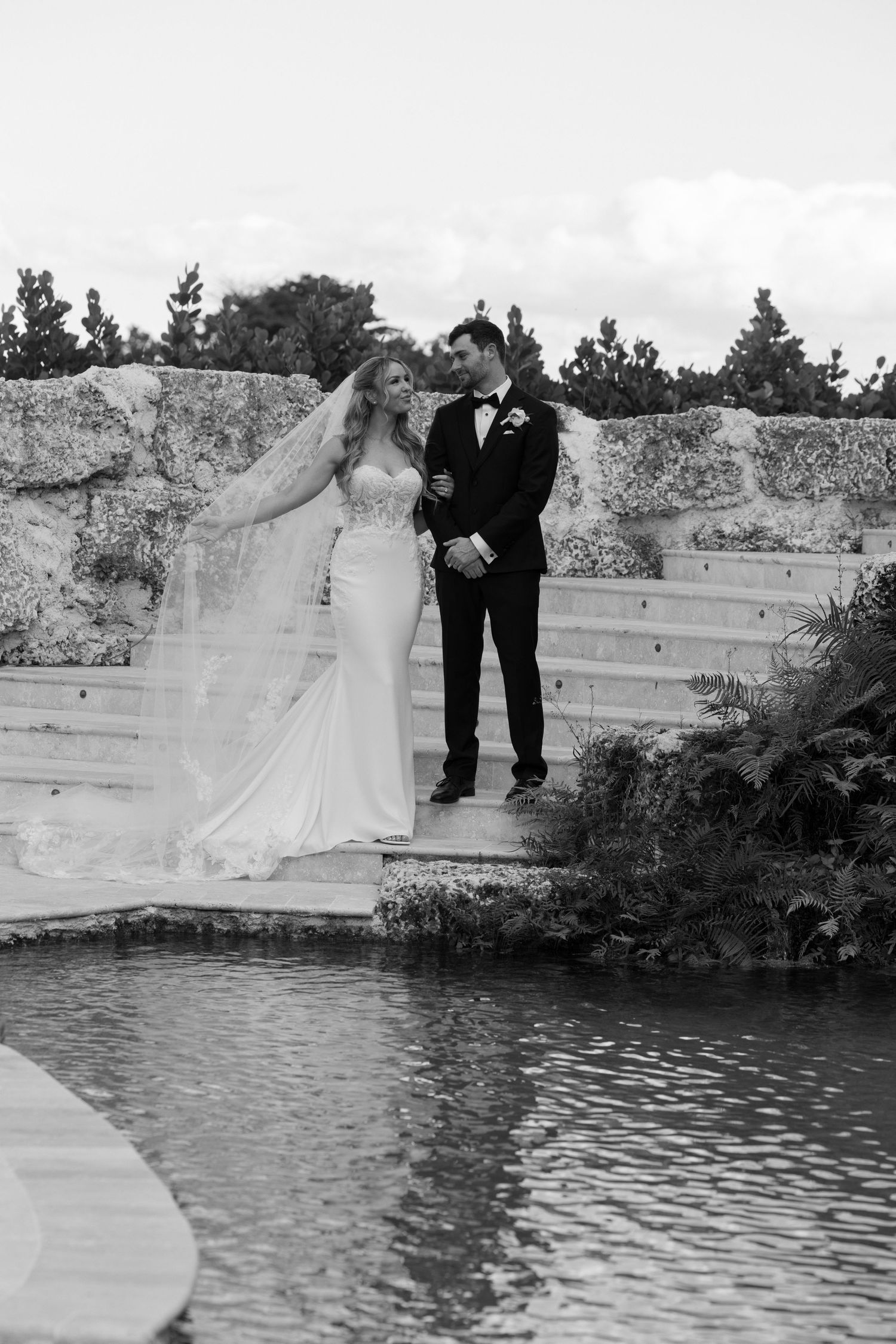 A couple in wedding attire stands on a stone wall overlooking a serene lake in a black and white photograph.