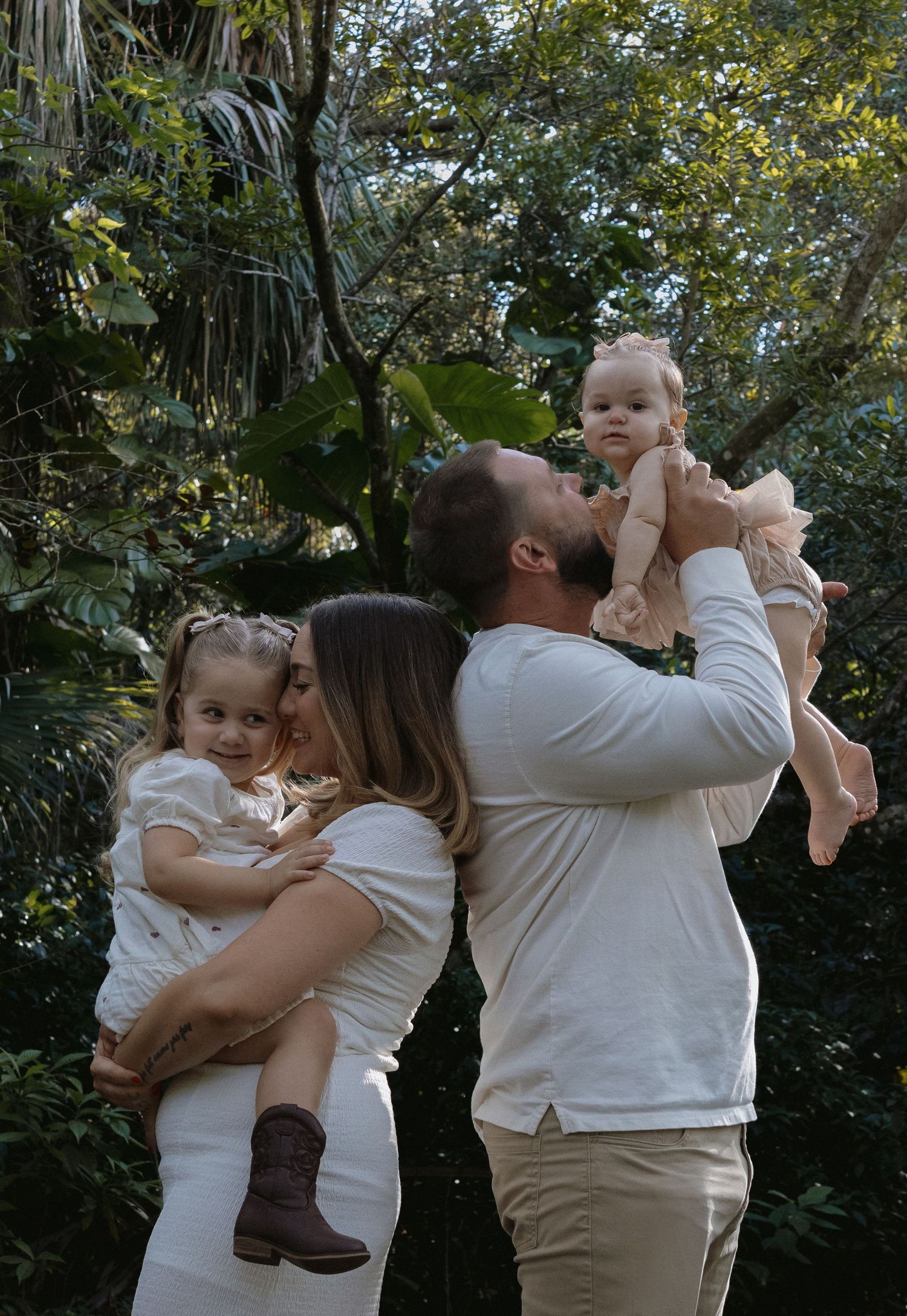 A family wearing white and beige clothing embraces and plays together in a lush outdoor garden setting.