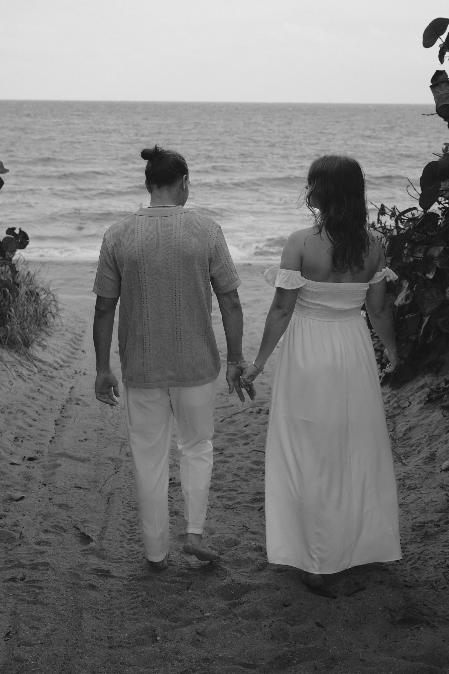 A romantic black and white beach scene showing a couple holding hands while walking on sandy path towards the ocean.
