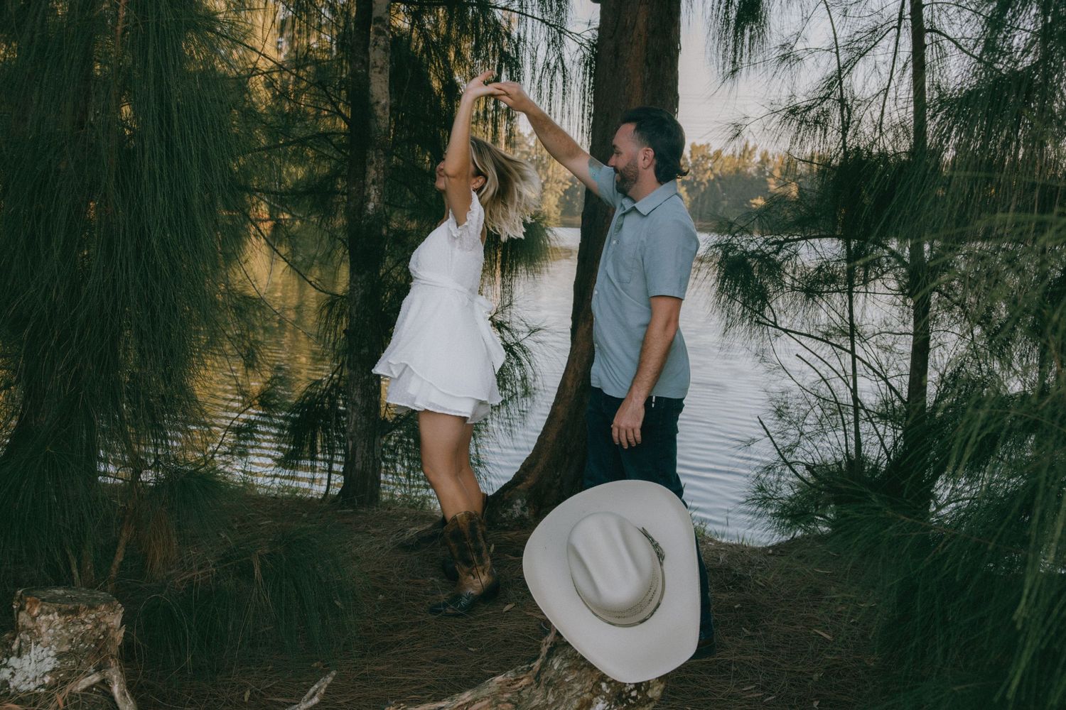 A romantic couple in casual attire shares a playful moment by a tranquil lake during sunset.