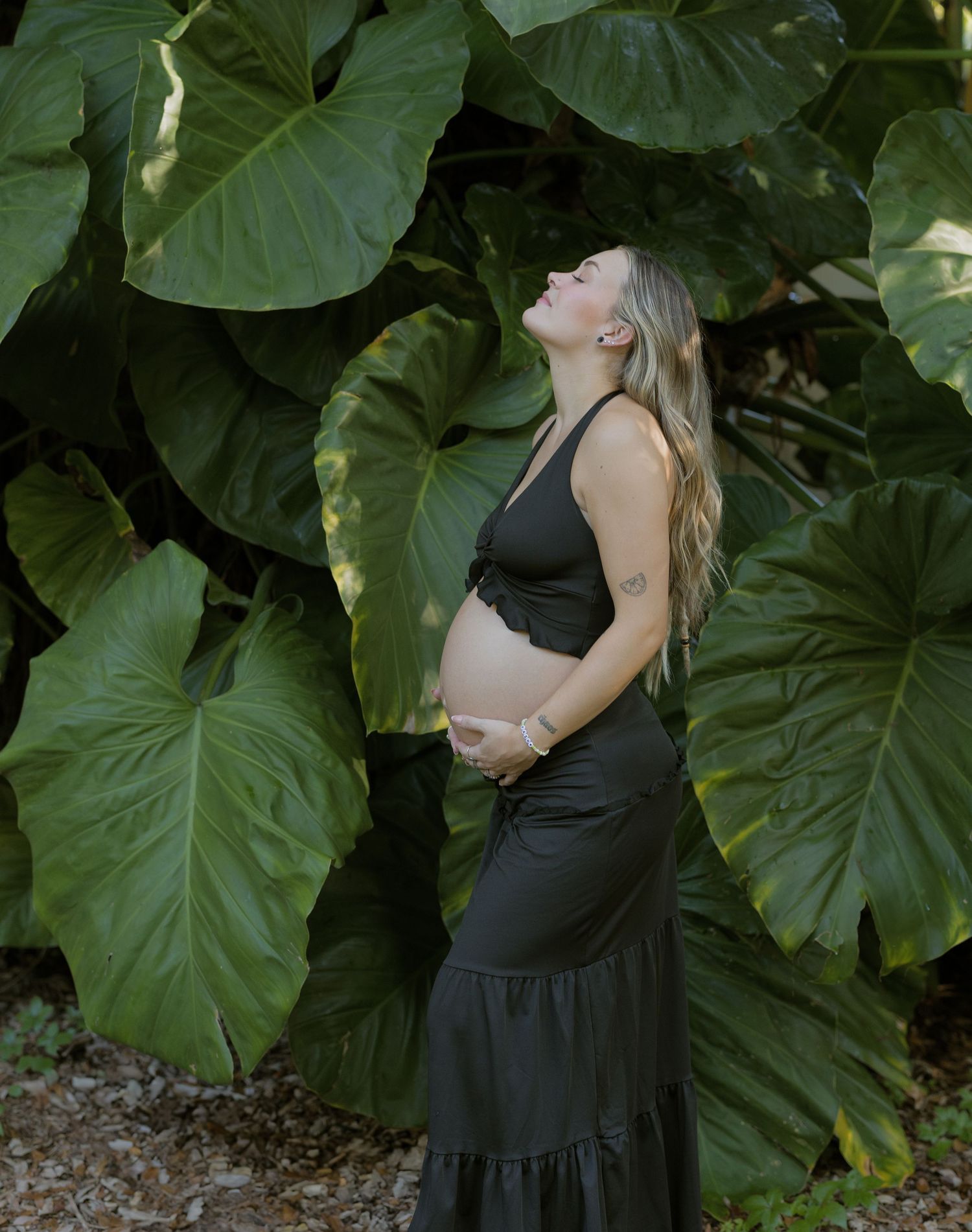 A pregnant person in dark clothing poses against large tropical elephant ear leaves in a garden setting.