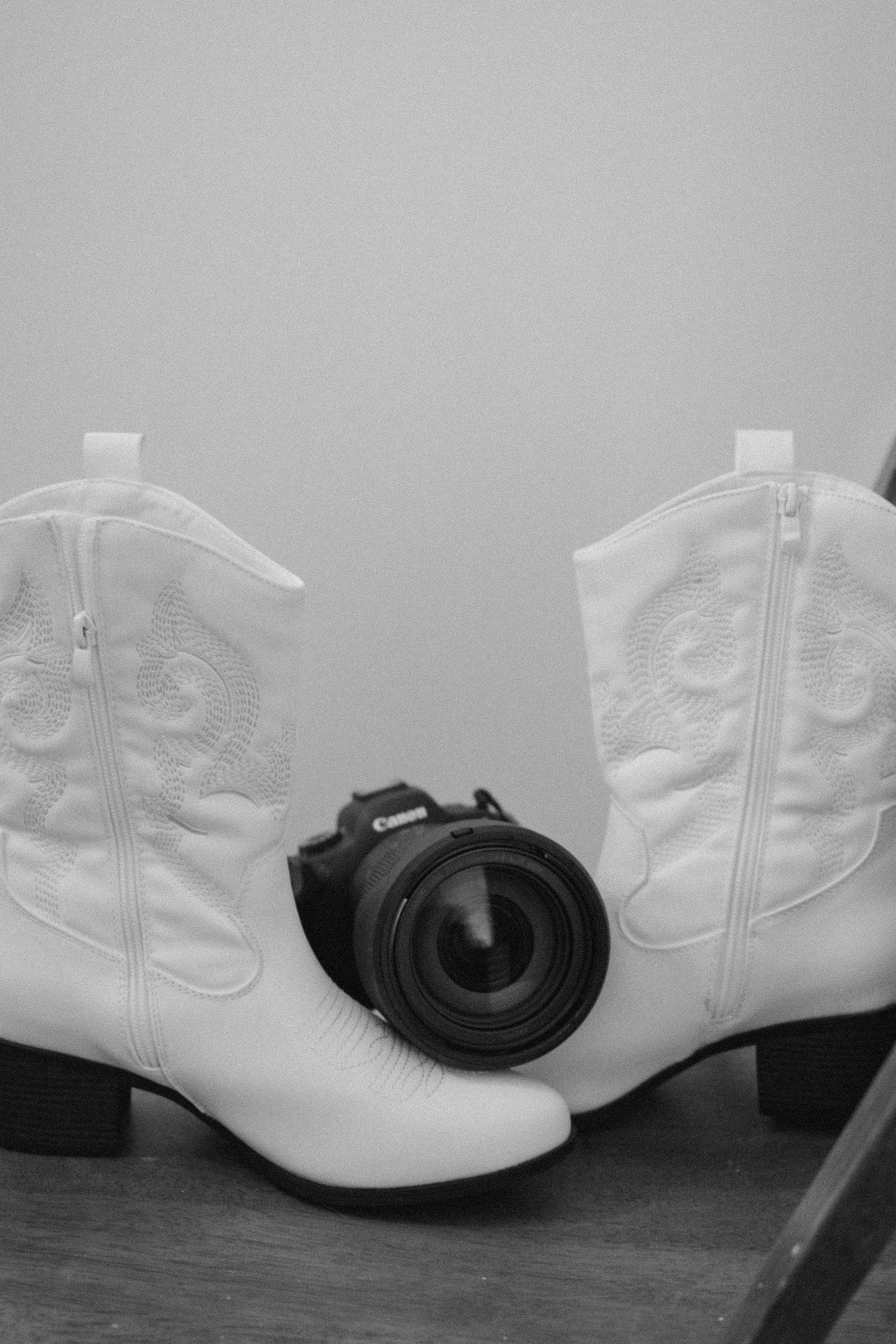 Black and white photo of white western cowboy boots with a black camera lens positioned between them on wooden floor.